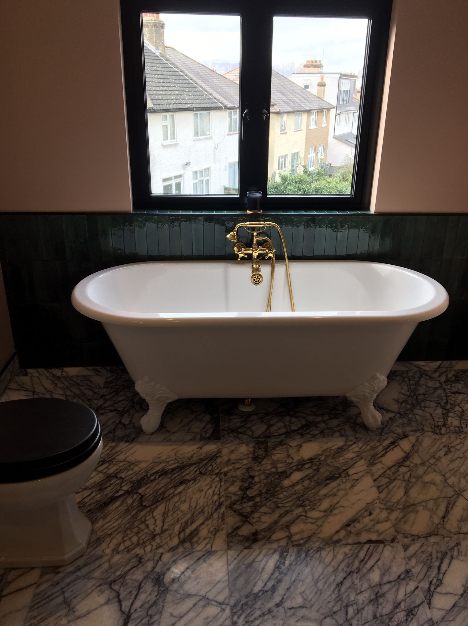 Biała wanna wolnostojąca na marmurowej podłodze, z czarną armaturą i złotą baterią, widok z okna na zabudowę miejską, ściany w kolorze pudrowego różu.