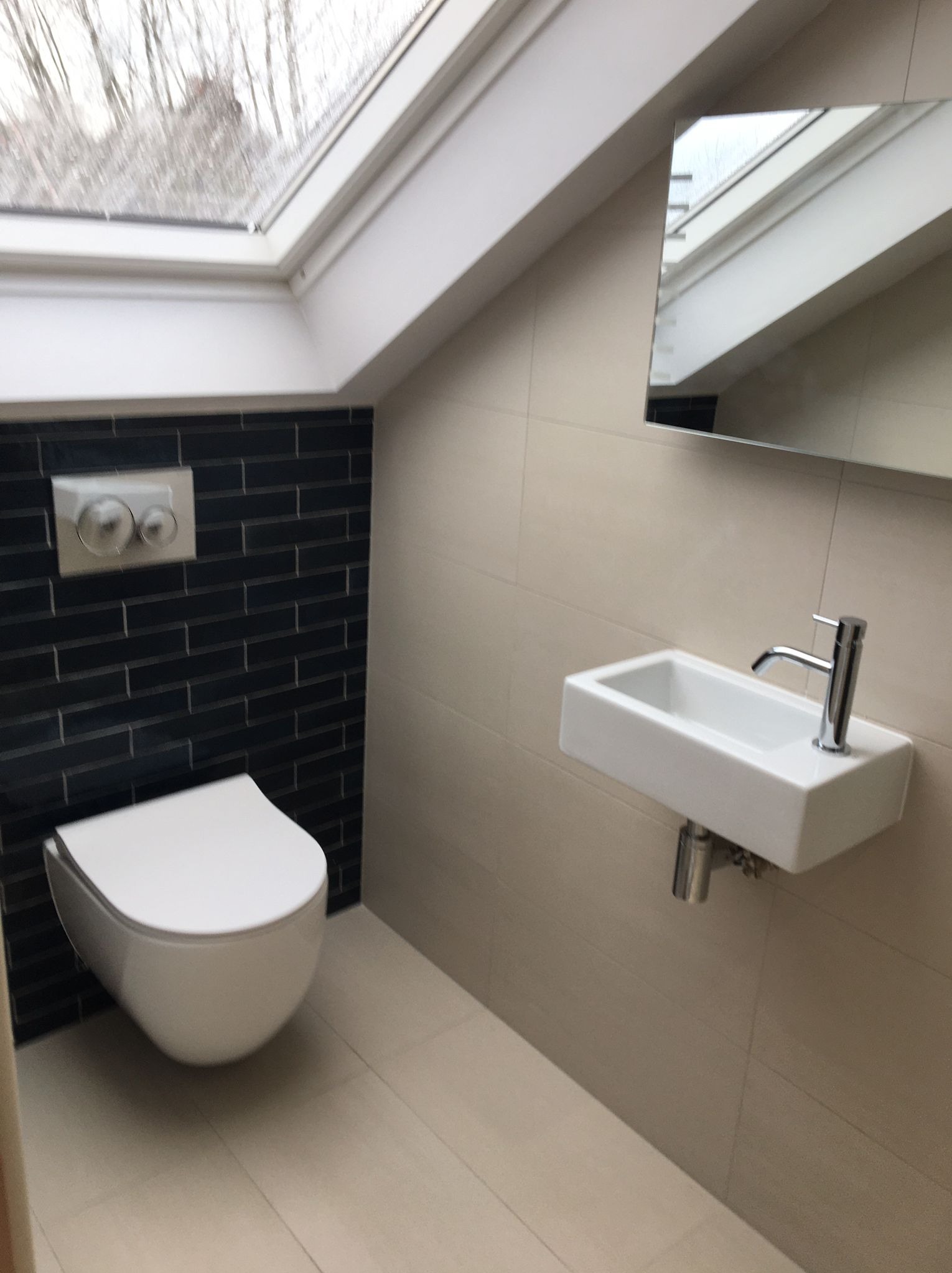 Nowoczesna łazienka na poddaszu z oknem dachowym: biała ceramika sanitarna (toaleta i umywalka), czarne płytki na ścianie, beżowe płytki podłogowe i ścienne, minimalistyczne lustro.