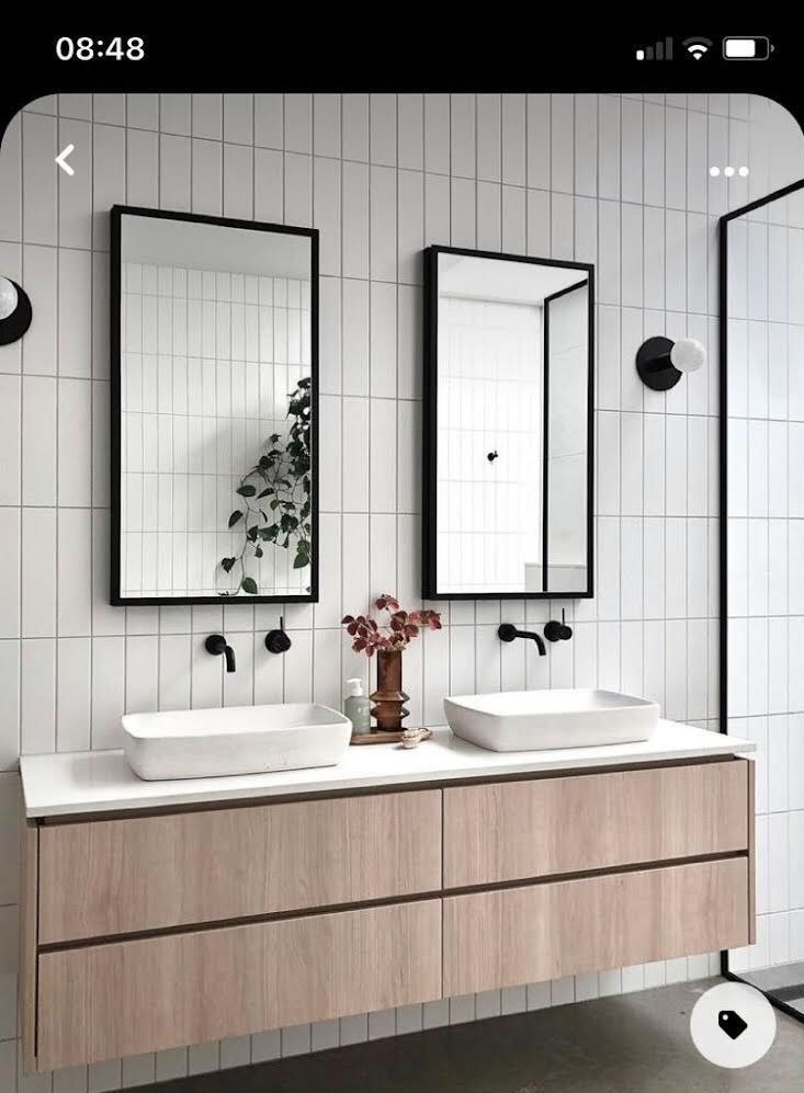 Nowoczesna łazienka z dwoma prostokątnymi umywalkami nablatowymi, czarnymi bateriami i lustrami w czarnych ramach na tle białych, pionowo ułożonych płytek. Na blacie stoi wazon z suszonymi kwiatami...