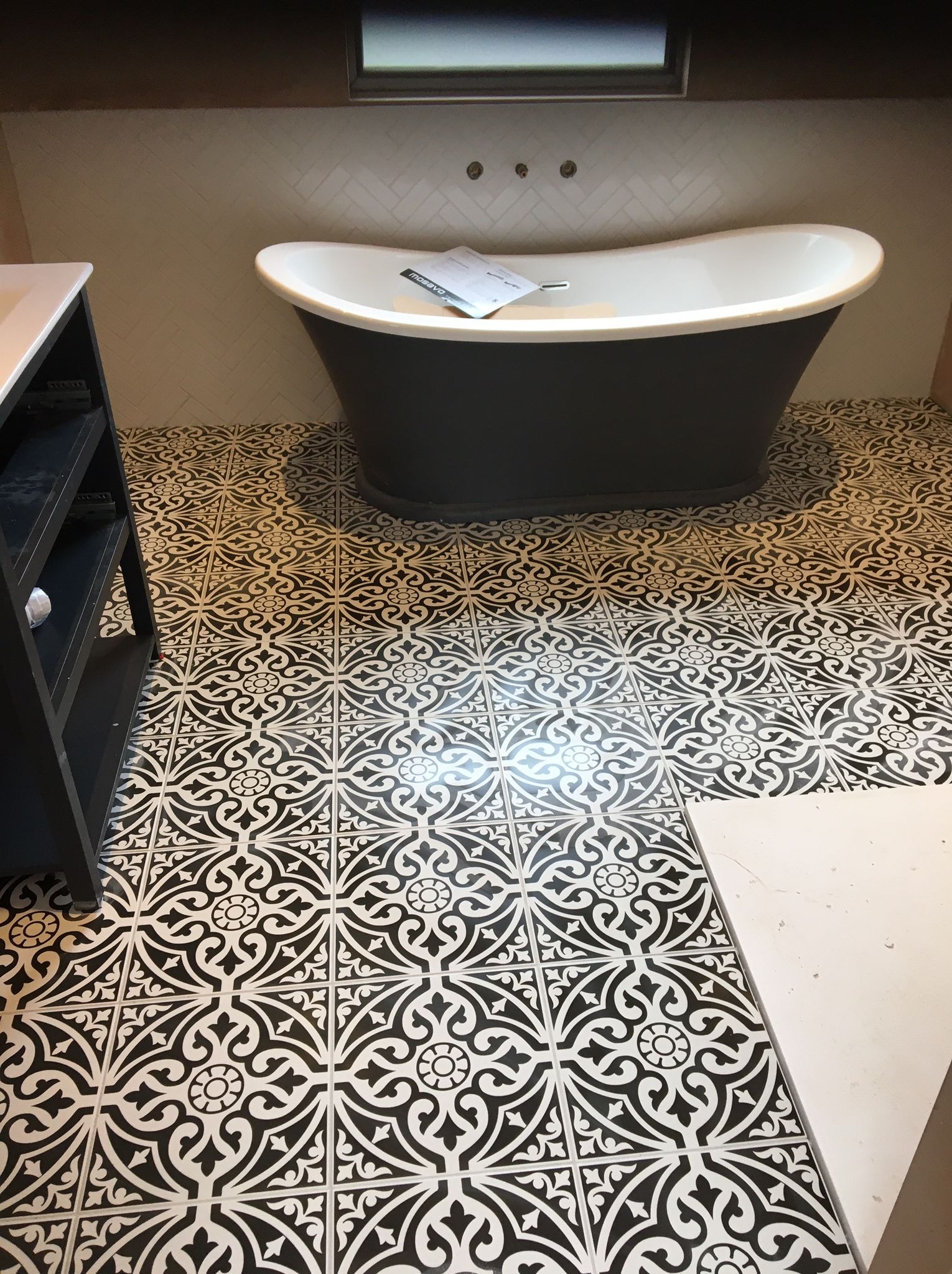 Czarno-biała łazienka z wolnostojącą wanną na tle wzorzystej podłogi i ściany wyłożonej białymi płytkami w jodełkę.