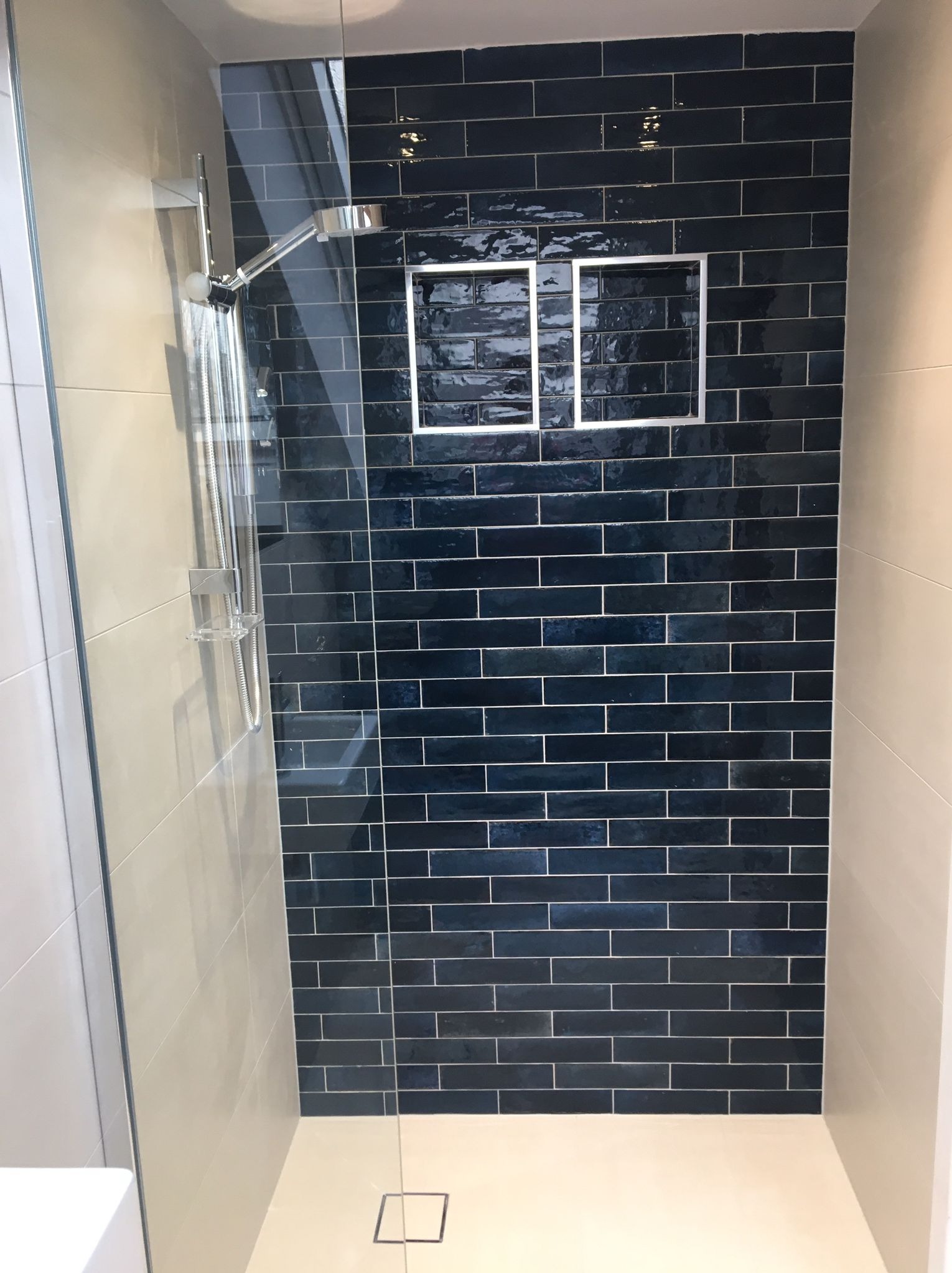 Kabina prysznicowa z ciemnoniebieskimi, błyszczącymi płytkami ułożonymi w cegiełkę, wnęka z podświetleniem, chromowana armatura i odpływ liniowy w podłodze.