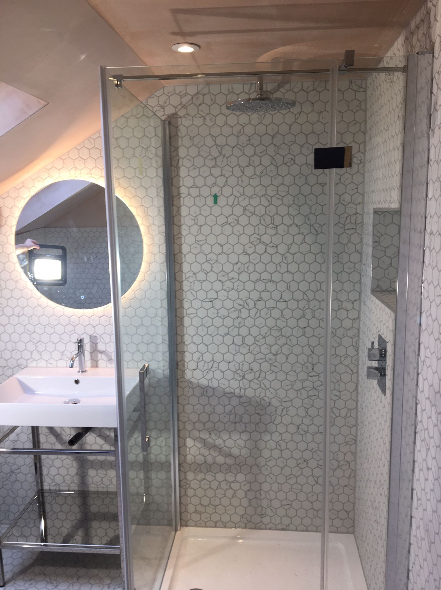 Nowoczesna łazienka na poddaszu z kabiną prysznicową ze szklanymi drzwiami, umywalką na metalowym stelażu i okrągłym lustrem z podświetleniem LED, wykończona białymi heksagonalnymi płytkami...