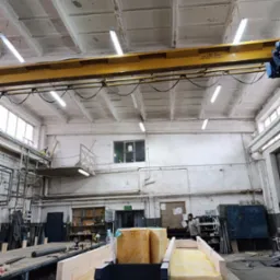 Prefabrykowane elementy konstrukcji dachu i ścian z izolacją w hali produkcyjnej. Widoczny surowy strop i oświetlenie. W tle pracownik w masce. Montaż na placu budowy.