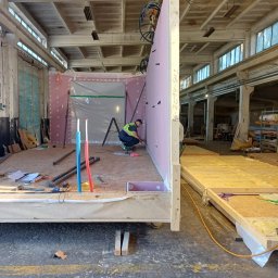 DOM DOBRY Roman K&K Sp. z o.o. - Montaż prefabrykowanych elementów domu modułowego w hali produkcyjnej. Widoczny pracownik w kamizelce, ściany z izolacją i fragment konstrukcji podłogi.