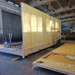 DOM DOBRY Roman K&K Sp. z o.o. - Prefabrykowane elementy konstrukcji modułowej domu z izolacją, podnoszone suwnicą w hali produkcyjnej. Widoczny pracownik i fragmenty innych modułów.