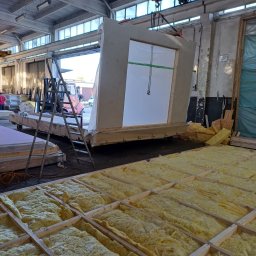 DOM DOBRY Roman K&K Sp. z o.o. - Prefabrykowany element ściany z oknem, podnoszony w hali produkcyjnej; widoczne rusztowanie, izolacja i suwnica z łańcuchem.