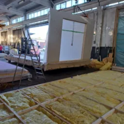 Prefabrykowany element ściany z oknem, podnoszony w hali produkcyjnej; widoczne rusztowanie, izolacja i suwnica z łańcuchem.