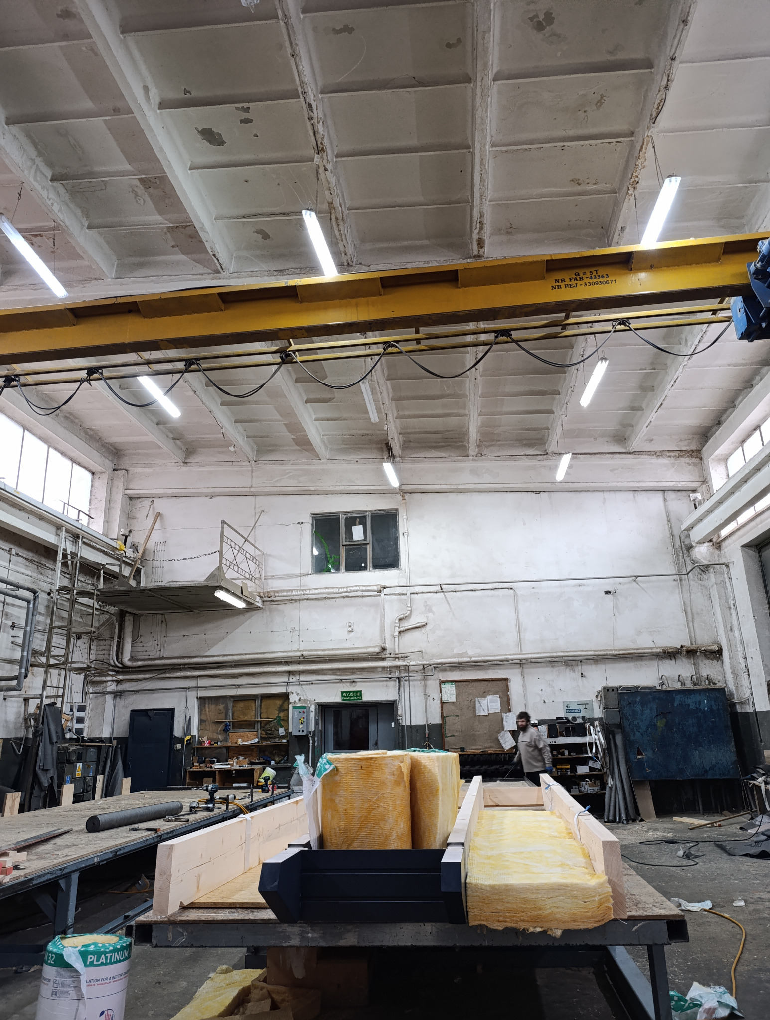 Prefabrykowane elementy konstrukcji dachu i ścian z izolacją w hali produkcyjnej. Widoczny surowy strop i oświetlenie. W tle pracownik w masce. Montaż na placu budowy.