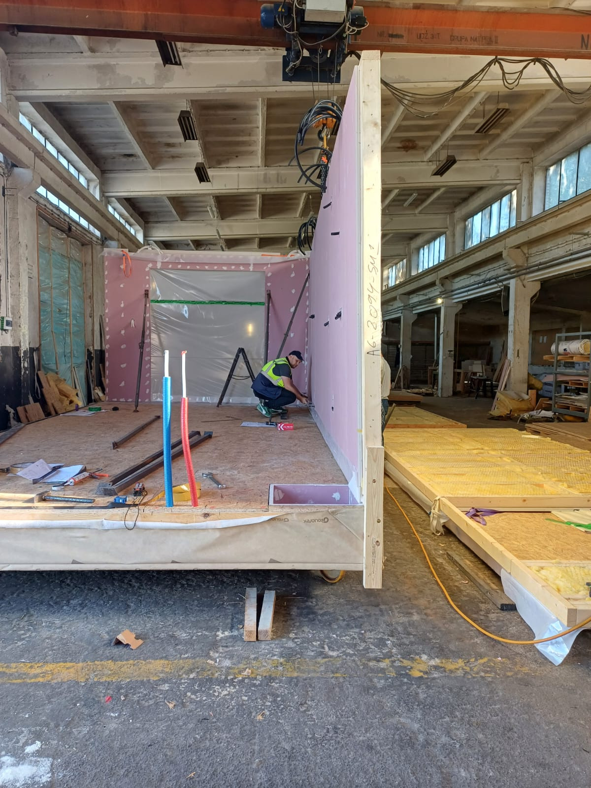 Montaż prefabrykowanych elementów domu modułowego w hali produkcyjnej. Widoczny pracownik w kamizelce, ściany z izolacją i fragment konstrukcji podłogi.