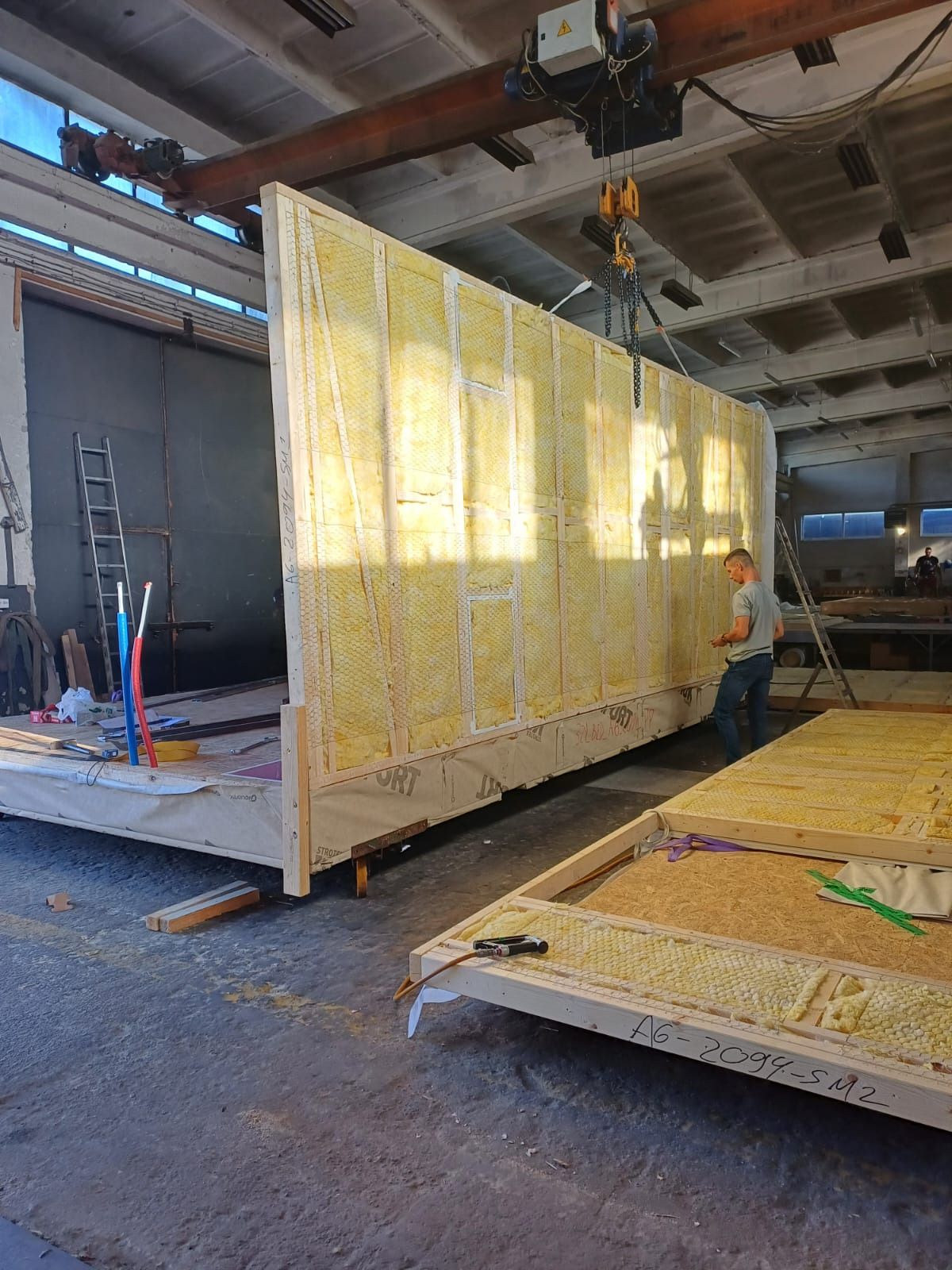 Prefabrykowane elementy konstrukcji modułowej domu z izolacją, podnoszone suwnicą w hali produkcyjnej. Widoczny pracownik i fragmenty innych modułów.