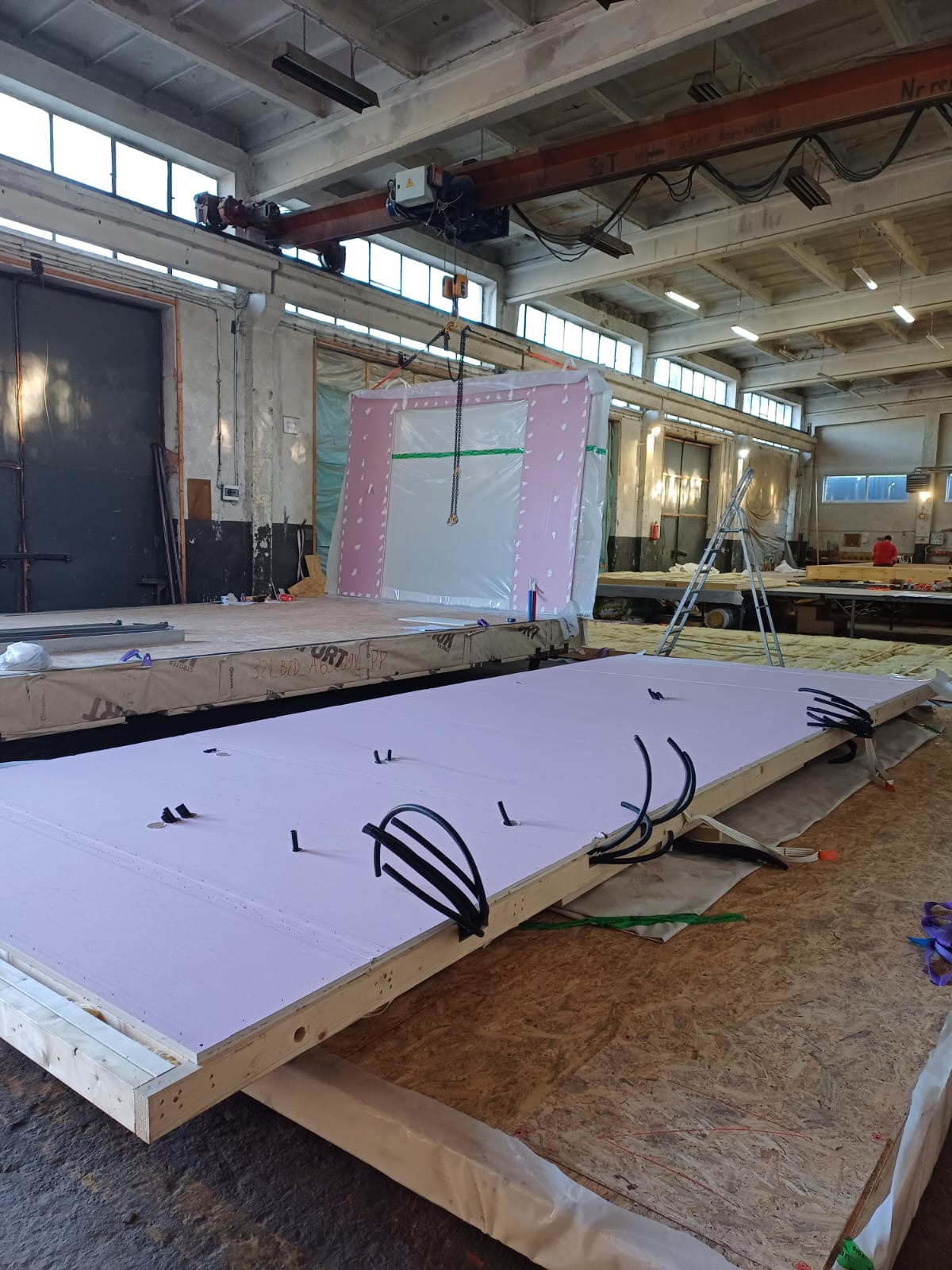 Hala produkcyjna: prefabrykowane elementy konstrukcji dachu i ścian z instalacjami, podnoszone przez suwnicę. Widoczne detale montażowe i izolacja.