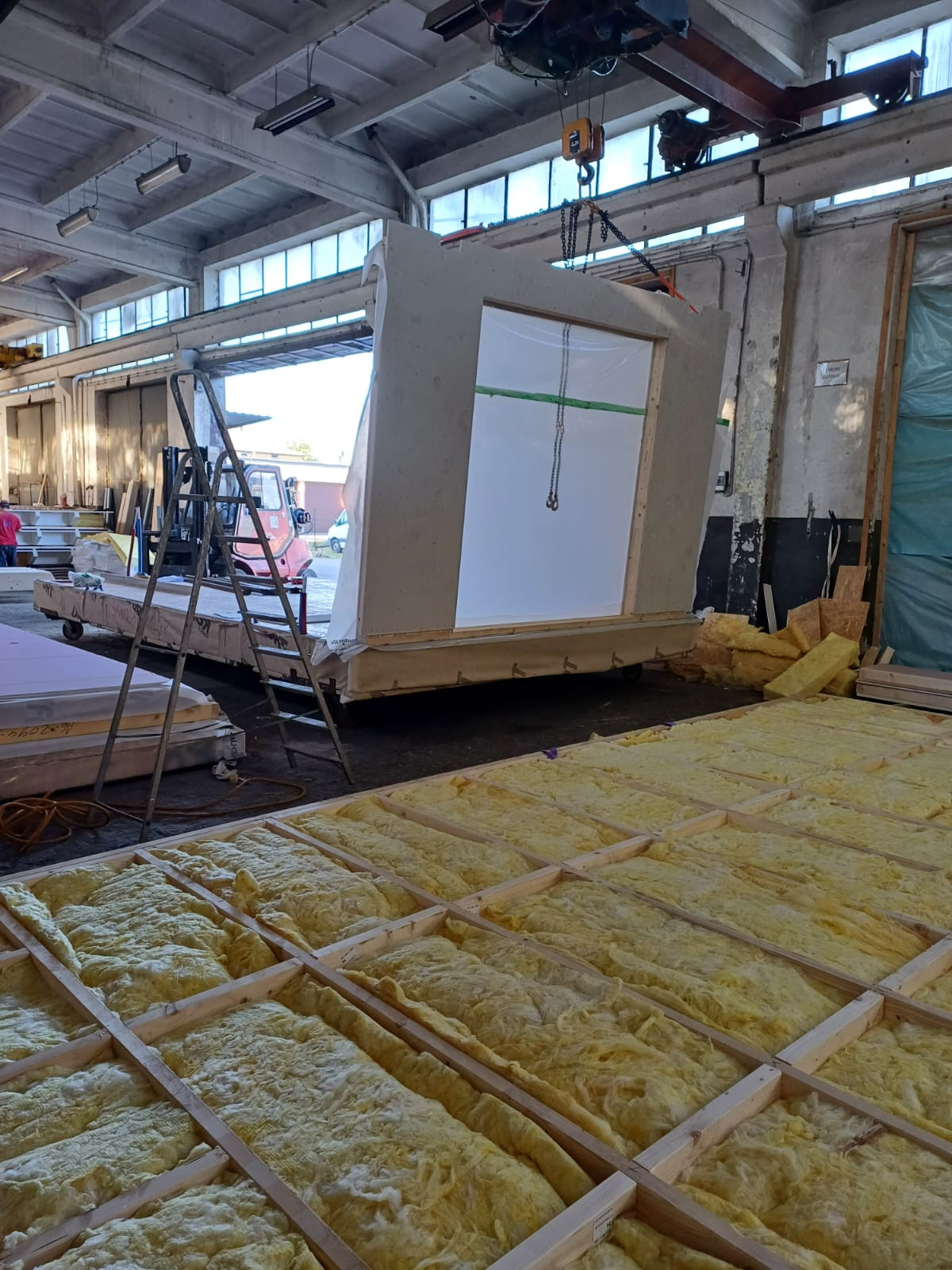 Prefabrykowany element ściany z oknem, podnoszony w hali produkcyjnej; widoczne rusztowanie, izolacja i suwnica z łańcuchem.