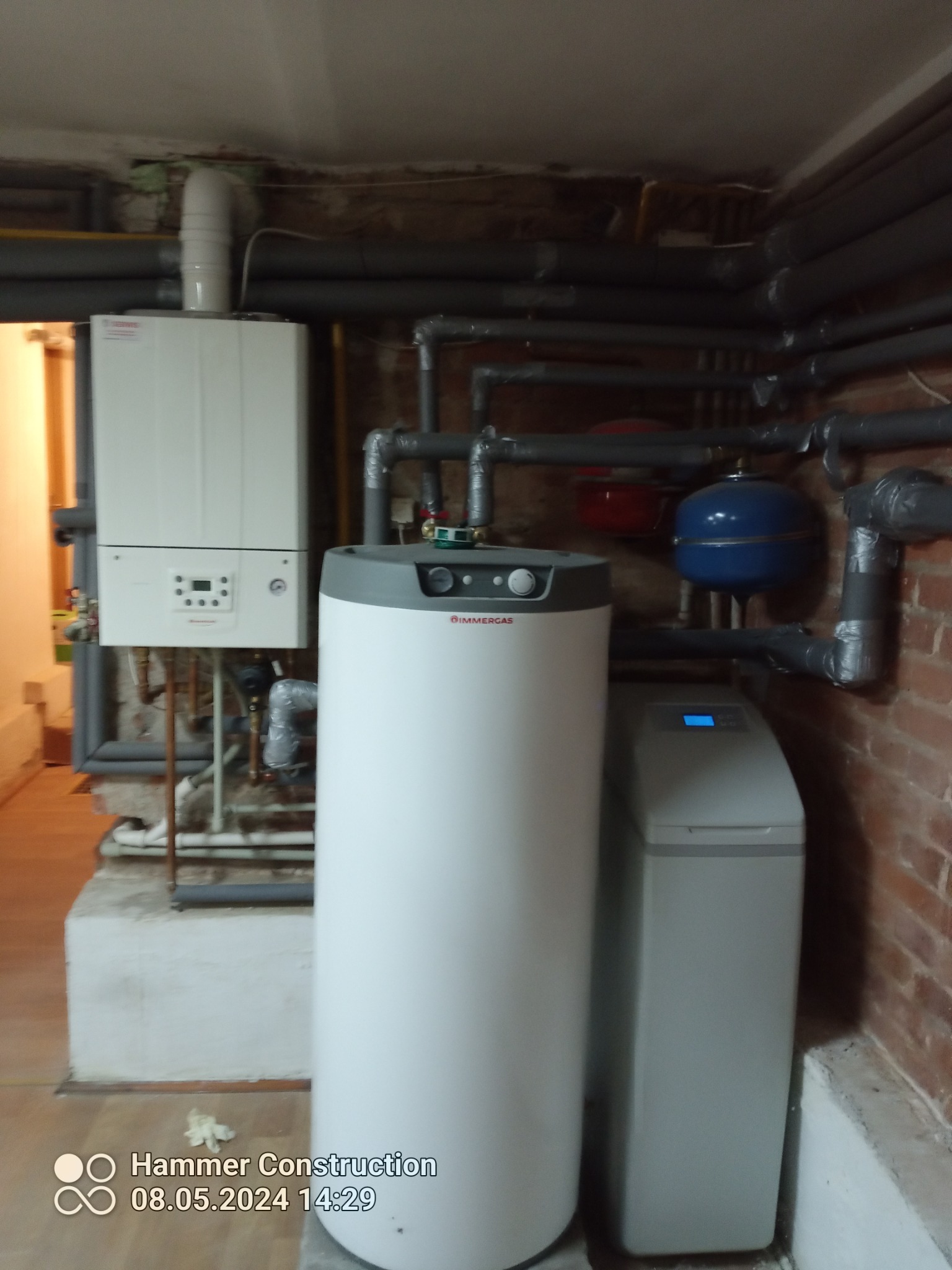 Nowoczesny piec gazowy Immergas, zasobnik ciepłej wody i zmiękczacz wody w kotłowni z widocznymi rurami grzewczymi i instalacją hydrauliczną.