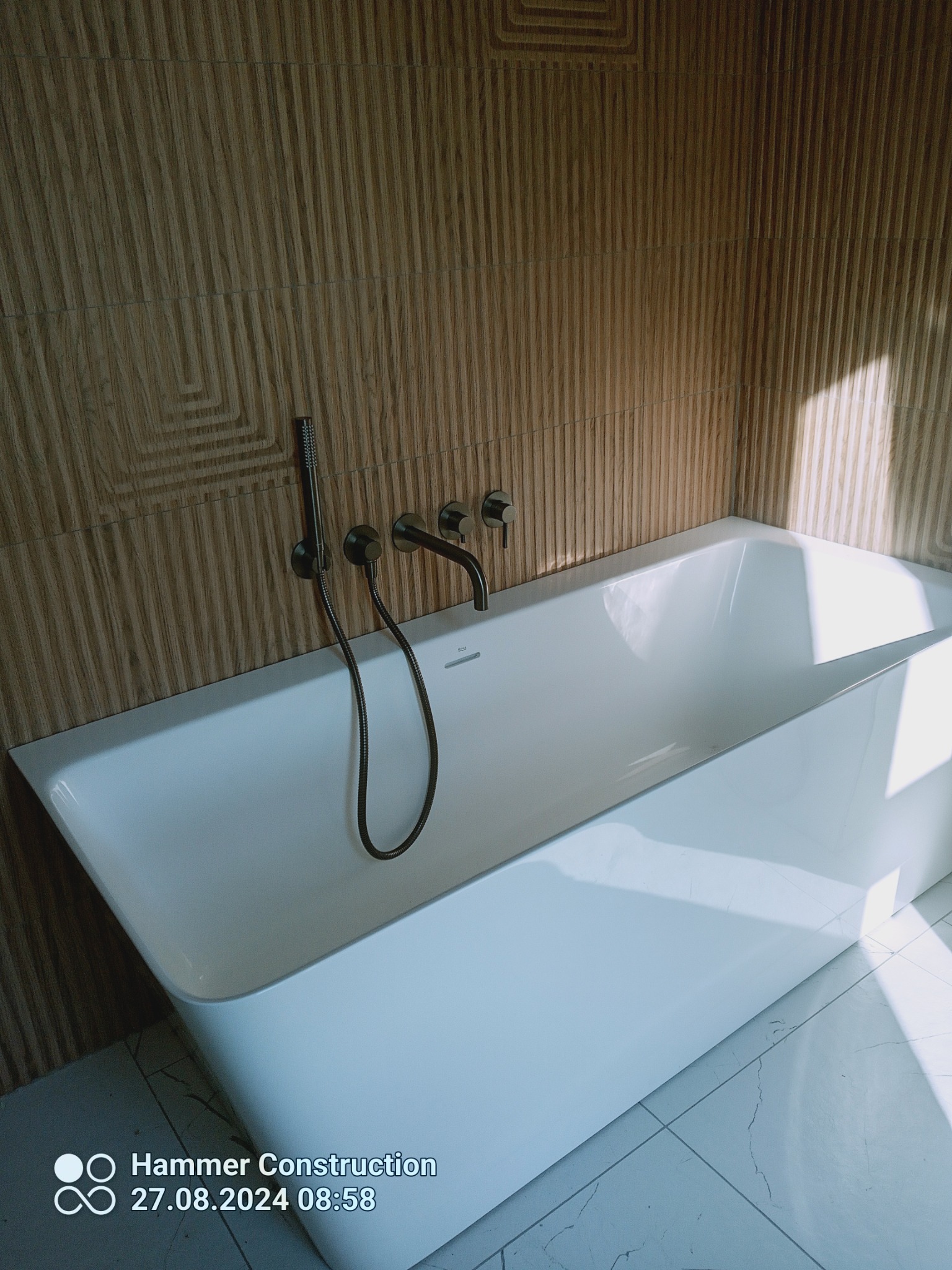 Nowoczesna, biała wanna z ciemną armaturą na tle ściany z drewnopodobnych paneli z geometrycznym wzorem, oświetlona naturalnym światłem.