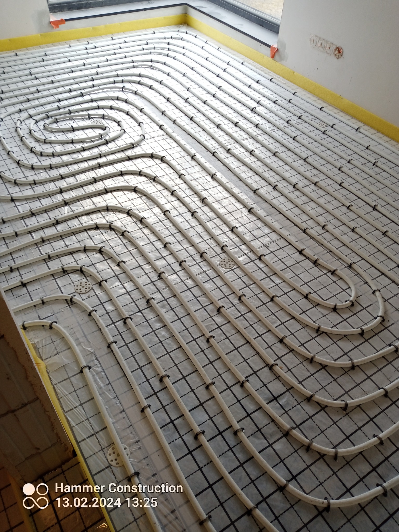 Białe rury ogrzewania podłogowego ułożone spiralnie i przymocowane do czarnej siatki na folii izolacyjnej, widok z góry w trakcie montażu.