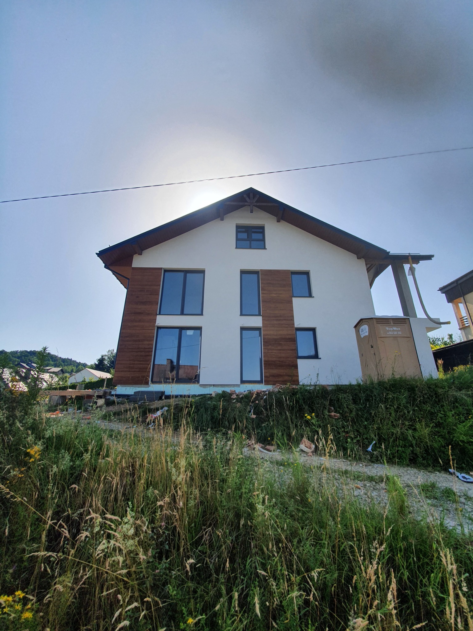 Nowoczesny dom jednorodzinny z białą elewacją i elementami dekoracyjnymi z paneli drewnopodobnych, widok z dołu przez wysoką trawę, w tle błękitne niebo z silnym słońcem.