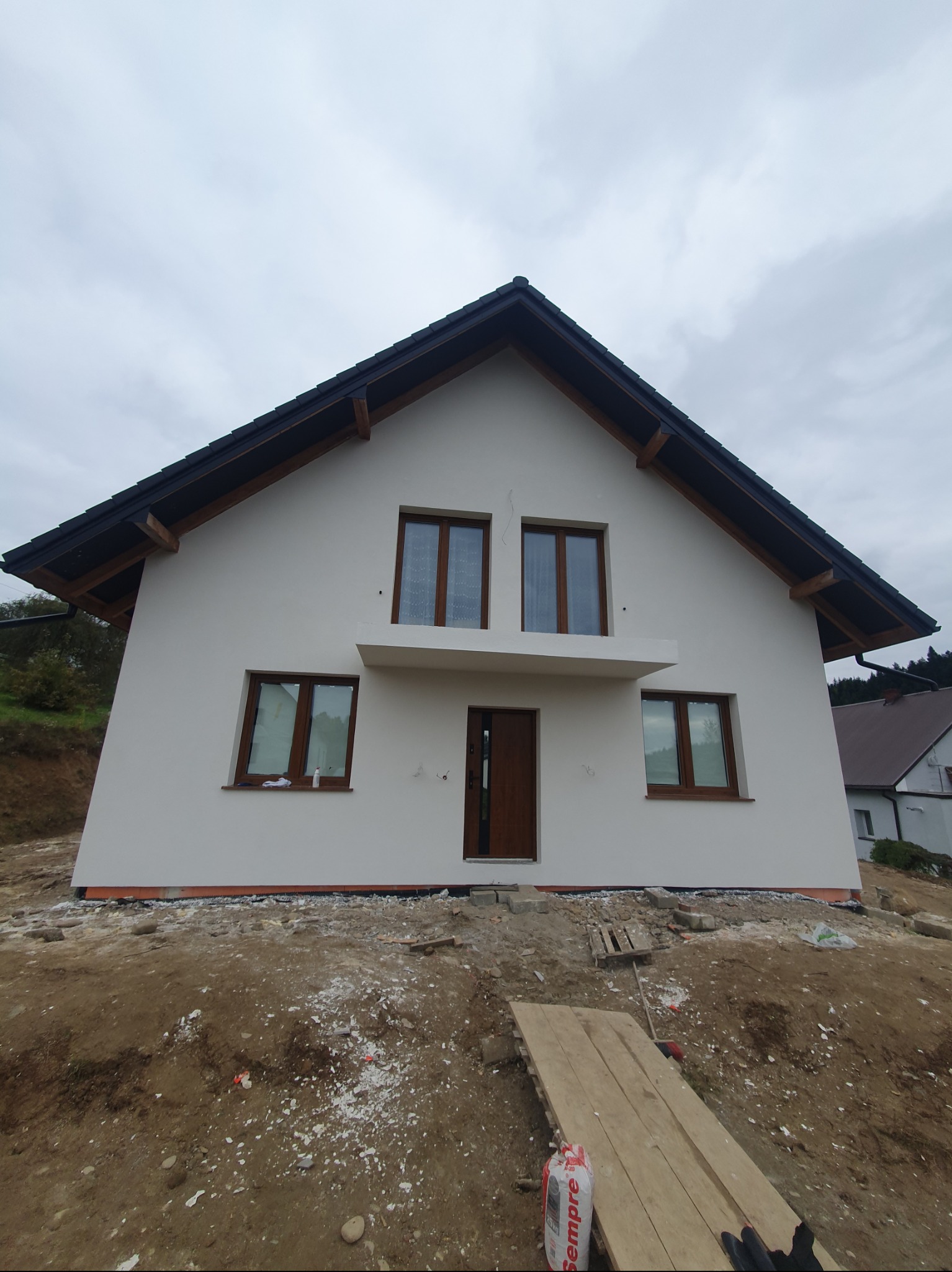 Nowoczesny dom jednorodzinny z jasną elewacją, brązowymi oknami i ciemnym dachem. Teren wokół domu w trakcie prac wykończeniowych, widoczne materiały budowlane.