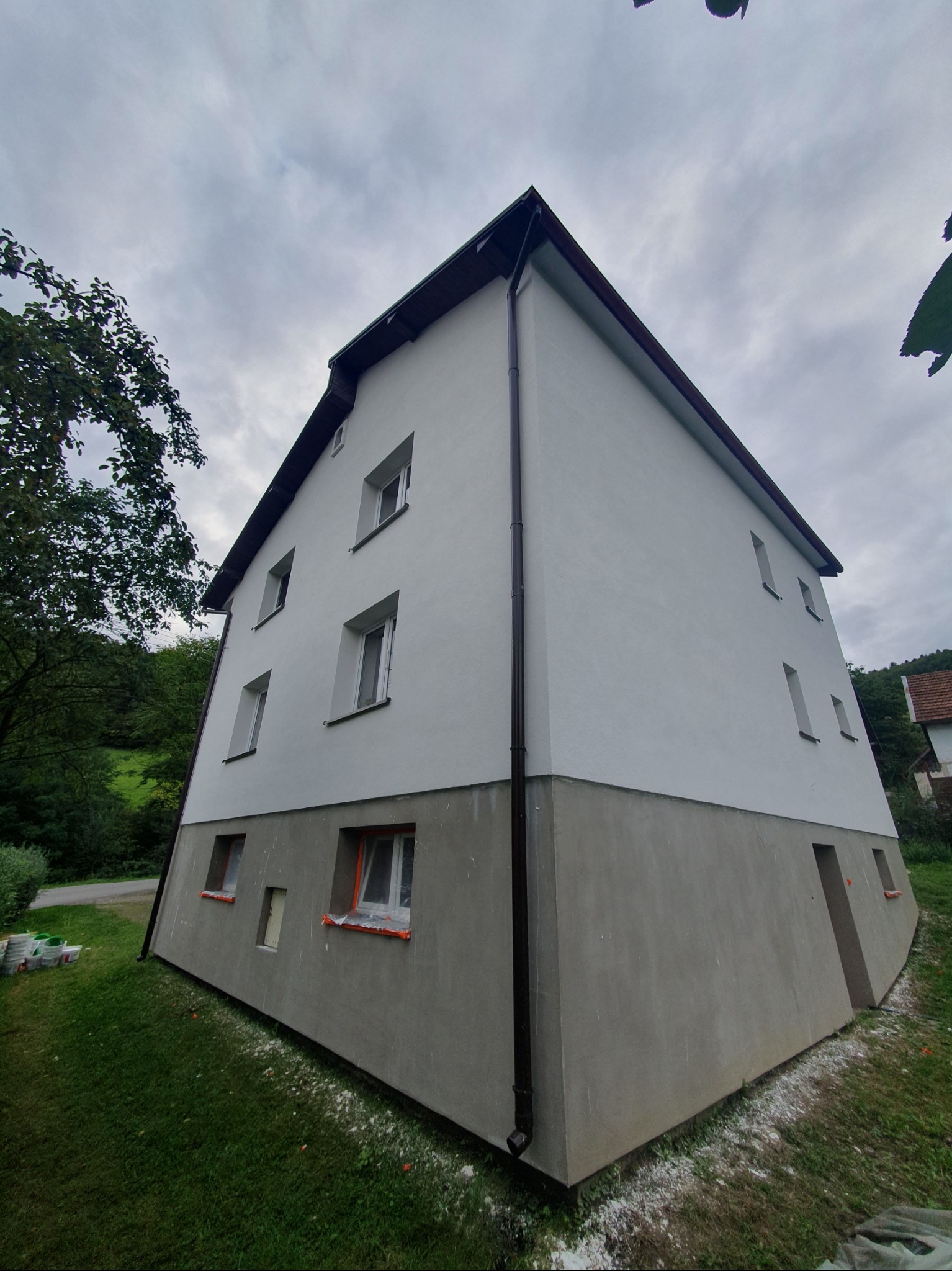 Wykończona elewacja dwukondygnacyjnego budynku z szarą dolną częścią i białą górną, widoczne okna z pomarańczowymi zabezpieczeniami, rynna i fragment zieleni.