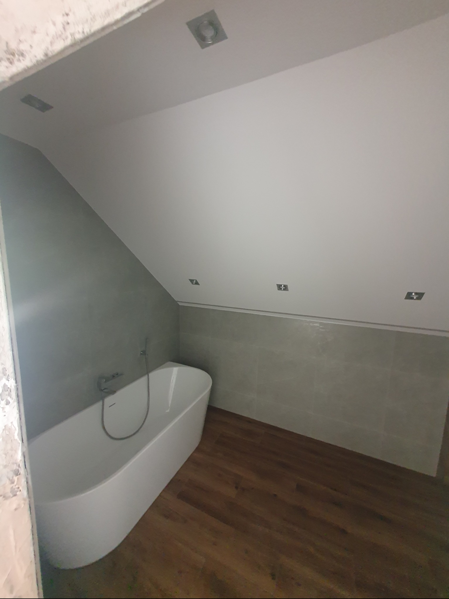 Nowoczesna łazienka na poddaszu z wolnostojącą wanną, szarą glazurą i drewnopodobną podłogą. Oświetlenie punktowe w suficie.