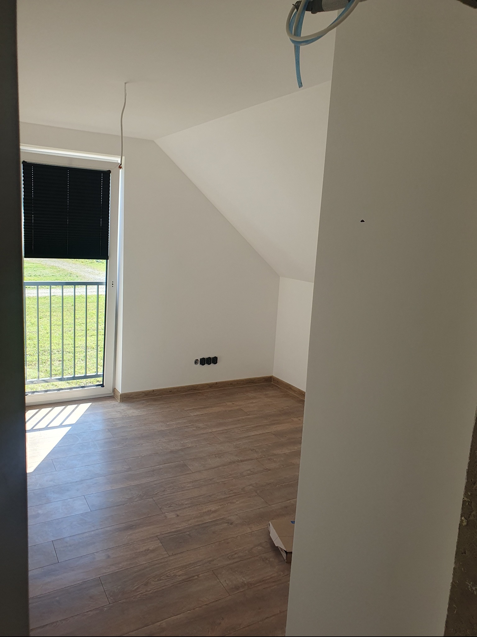 Poddasze z drewnopodobną podłogą, widok na zieleń przez okno z żaluzjami. Białe ściany, widoczne gniazdka elektryczne i rury instalacyjne pod sufitem.