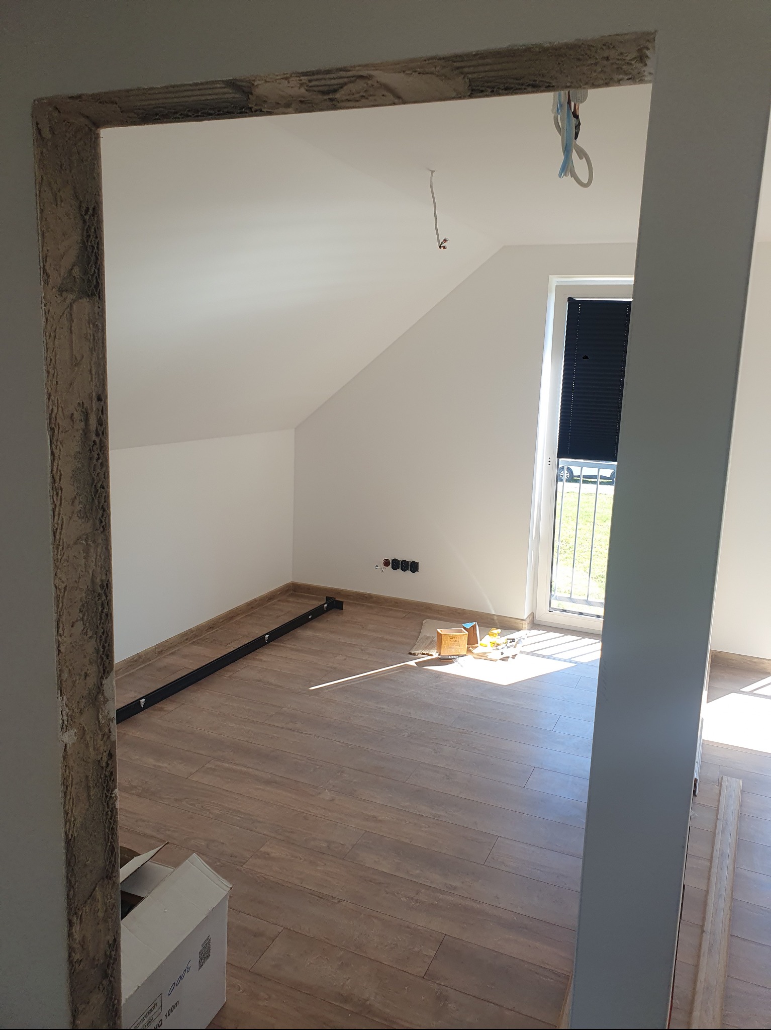 Podłoga z paneli imitujących drewno w jasnym pokoju na poddaszu, widok przez surową futrynę. Przy oknie balkonowym leżą narzędzia.