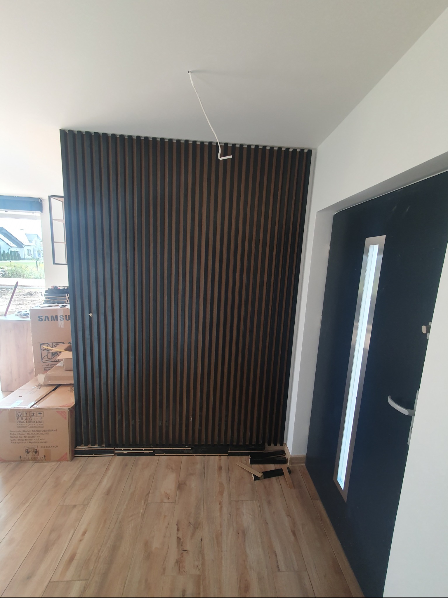Drewniane lamele na ścianie w przedpokoju, widoczne kartony z elektroniką, podłoga z paneli, czarne drzwi wejściowe z szybą. Nowoczesny design wnętrza.