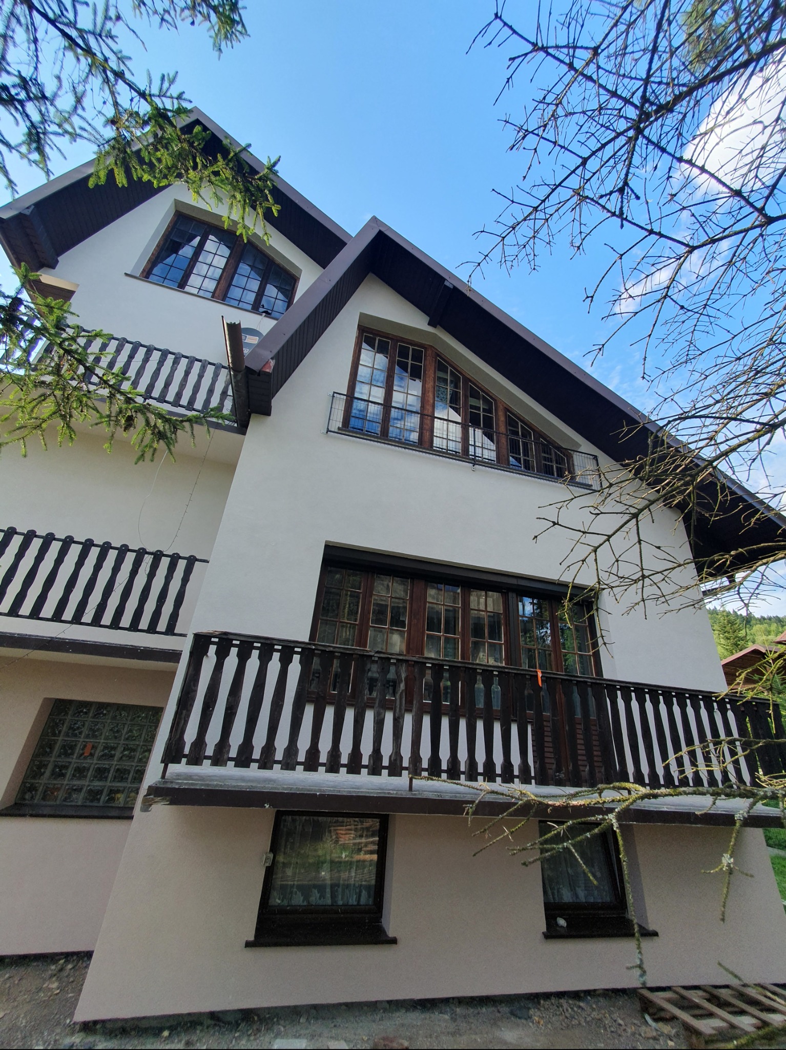 Dwupiętrowy dom z balkonami i oknami z drewnianą stolarką, widziany z dołu, z fragmentami drzew na pierwszym planie i błękitnym niebem w tle.