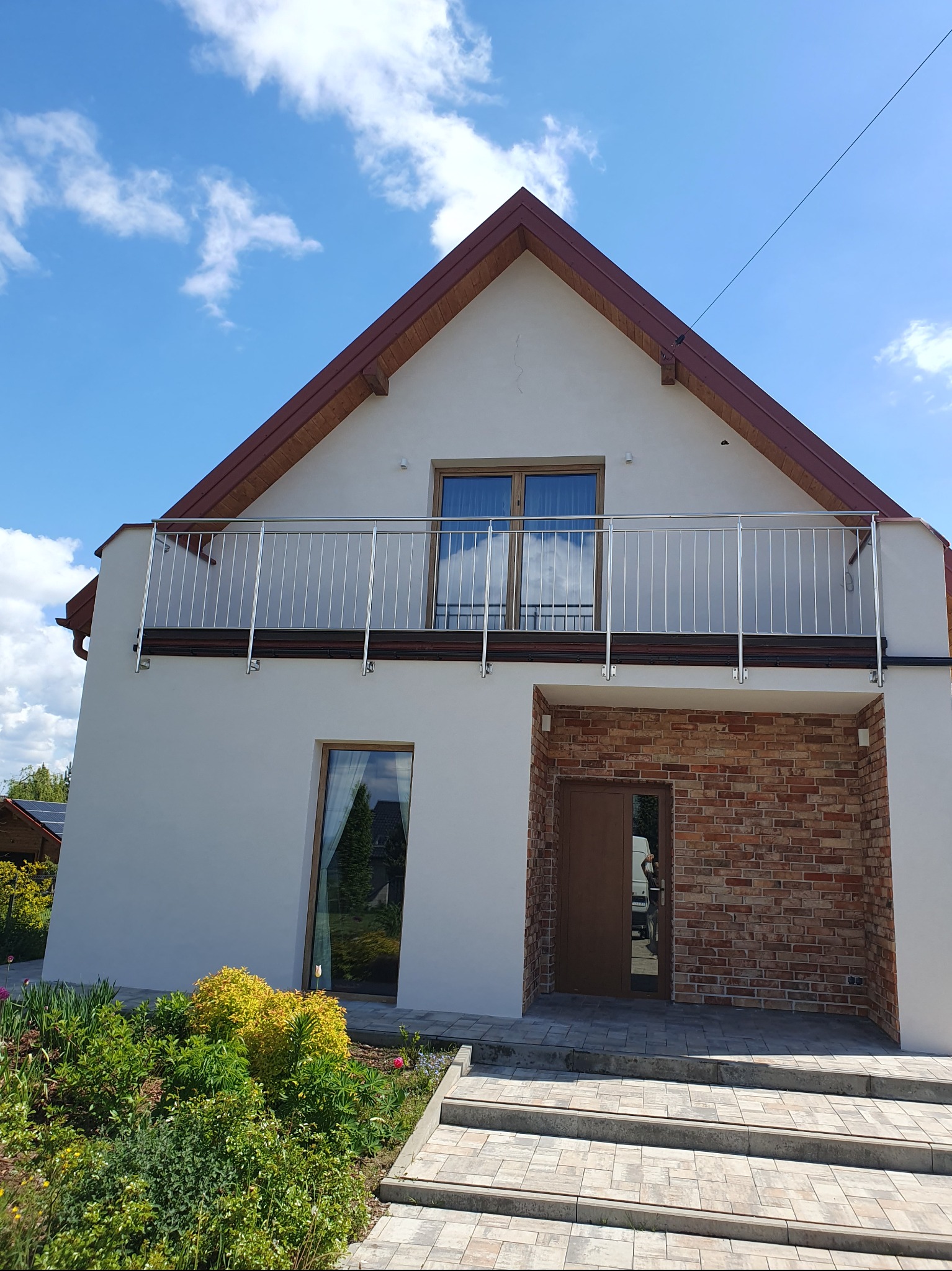 Dwukondygnacyjny dom z białymi ścianami, balkonem z metalową balustradą i wejściem wykończonym cegłą. Widoczne schody wejściowe i fragment ogrodu.