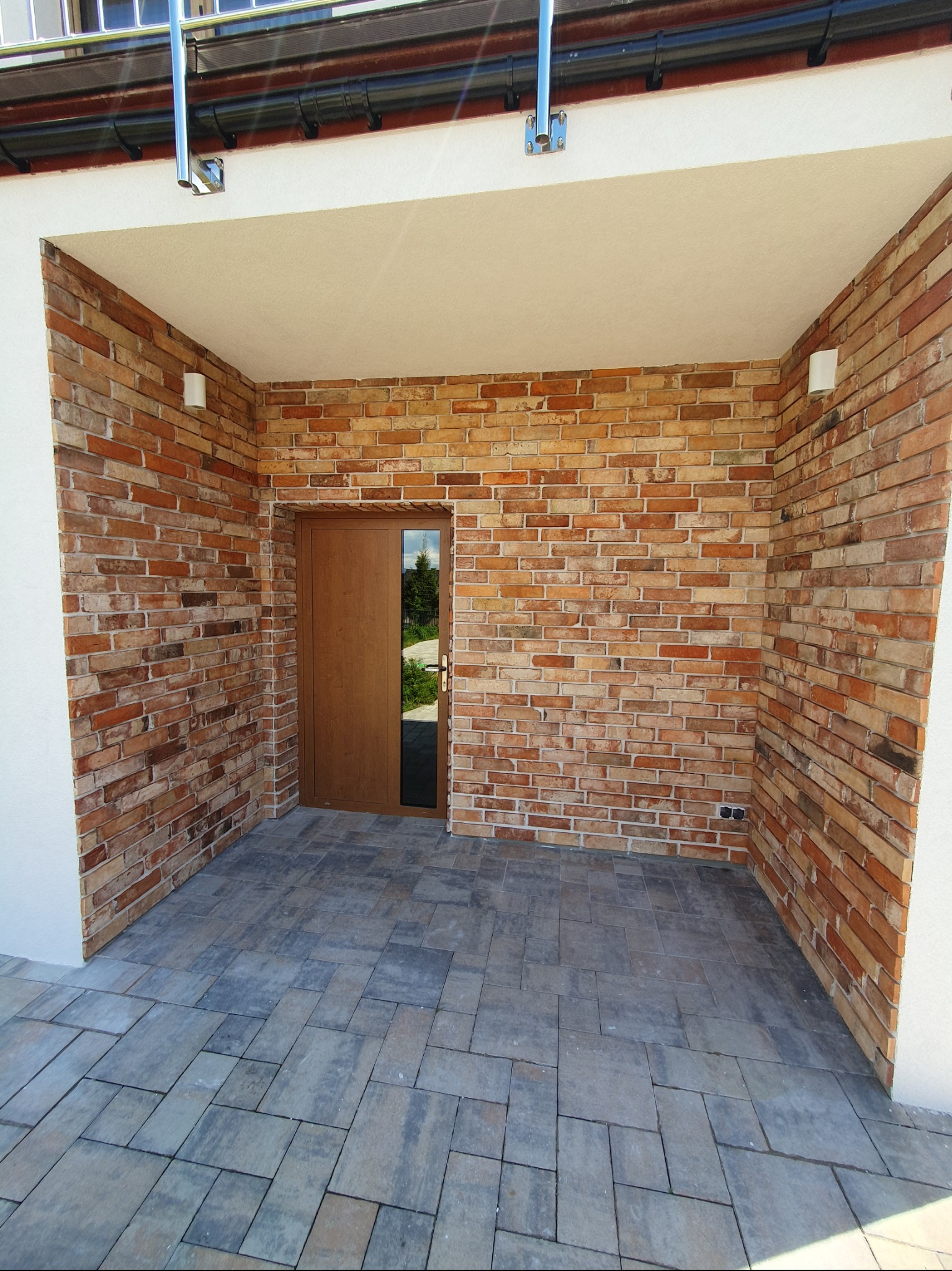 Zadaszone wejście do budynku z ceglaną ścianą i szarą kostką brukową. Widoczne oświetlenie zewnętrzne i brązowe drzwi z szybą. Detal wykończenia elewacji.