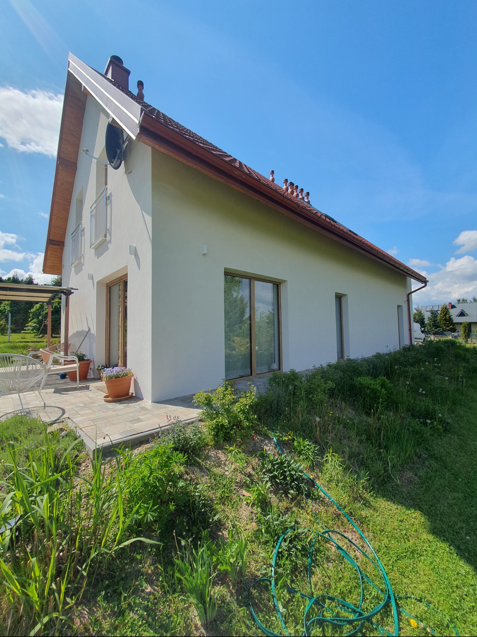 Biały dom jednorodzinny z brązowym dachem, widoczny z perspektywy ogrodu. Elewacja wykończona, okna z drewnianą stolarką. Na dachu liczne kominy.