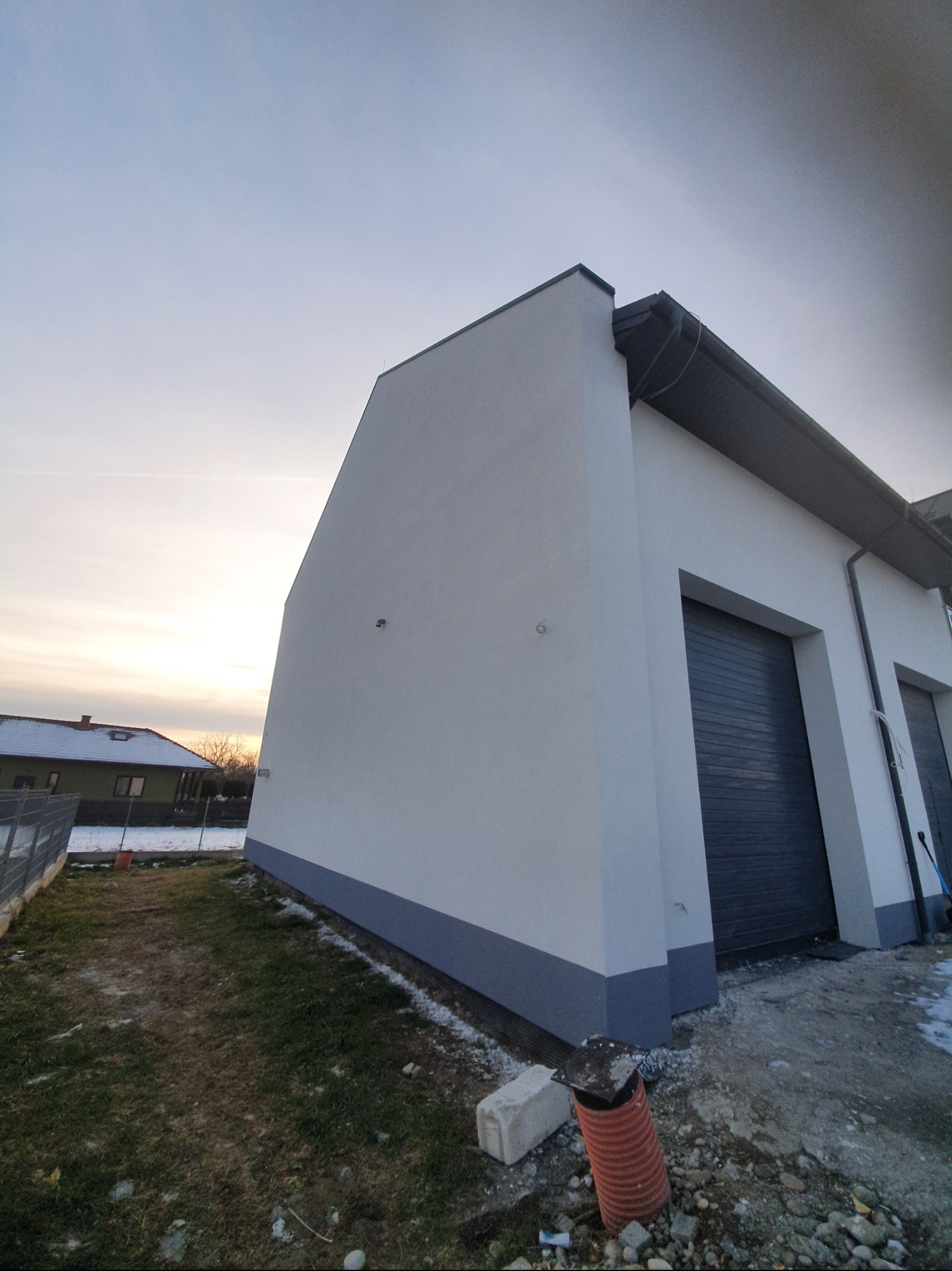 Biała elewacja budynku z szarą podmurówką i bramami garażowymi, widok z dołu, w tle dom z ośnieżonym dachem i zachodzące słońce.