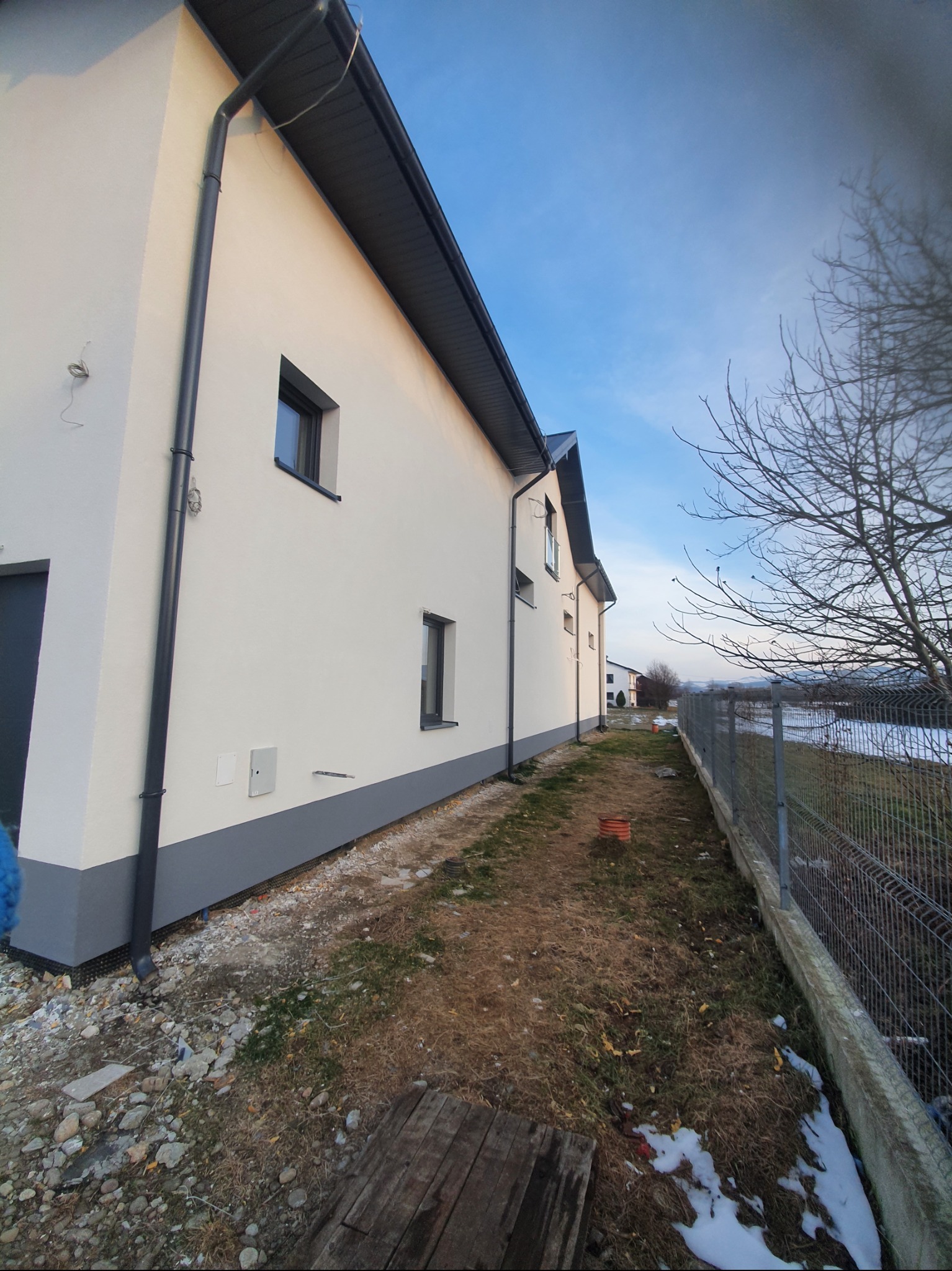 Świeżo otynkowana elewacja domu jednorodzinnego z szarym cokołem i orynnowaniem, widok z boku na tle błękitnego nieba i zimowego krajobrazu.