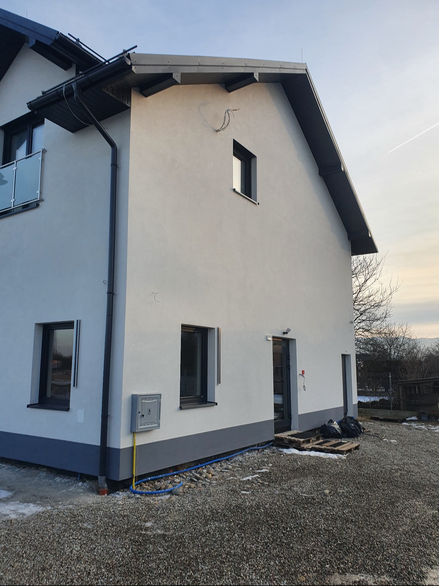 Nowoczesny dom jednorodzinny z jasną elewacją i ciemnymi detalami wykończeniowymi, widok z boku na tle błękitnego nieba i drzew. Widoczne okna i drzwi.