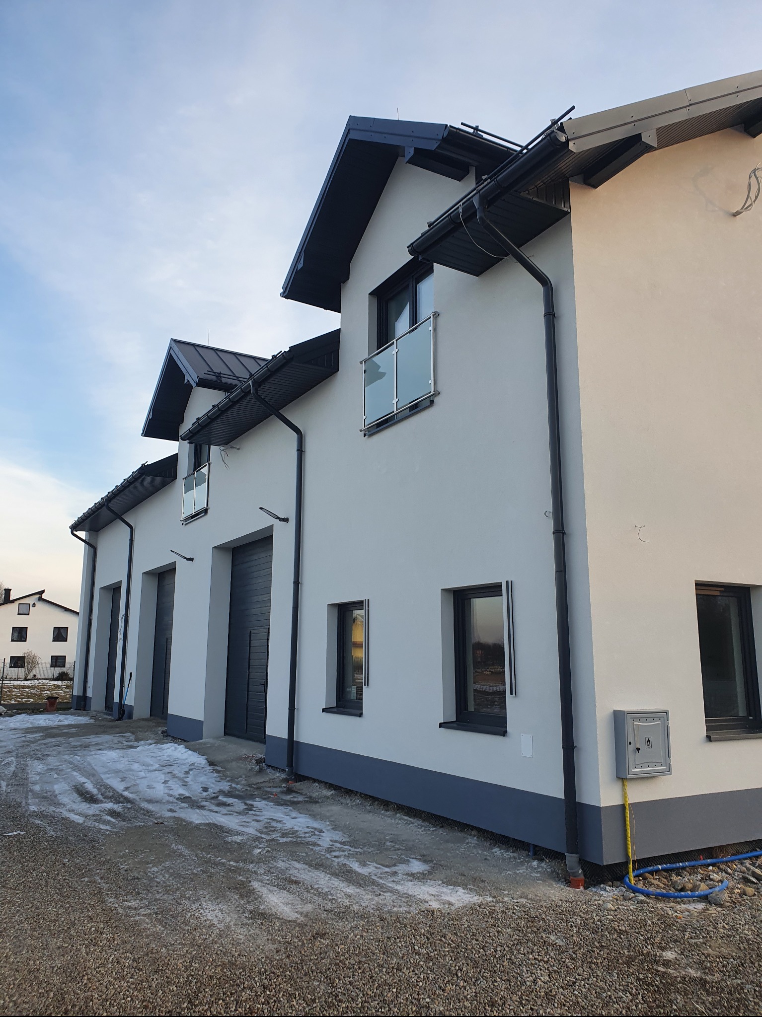 Nowoczesny budynek z szarą elewacją, czarnymi oknami i balkonami ze szklanymi balustradami. Widoczne rynny i instalacje przy budynku. Częściowo ośnieżony teren.