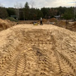 Praca zagęszczarką płytową na placu budowy, widoczne ślady opon na piasku, przygotowanie terenu pod budowę w otoczeniu drzew.