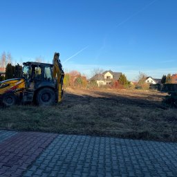 Mateusz Steć - Żółto-czarna koparko-ładowarka Case stoi na przygotowanym placu budowy z widokiem na domy jednorodzinne w tle, błękitne niebo.