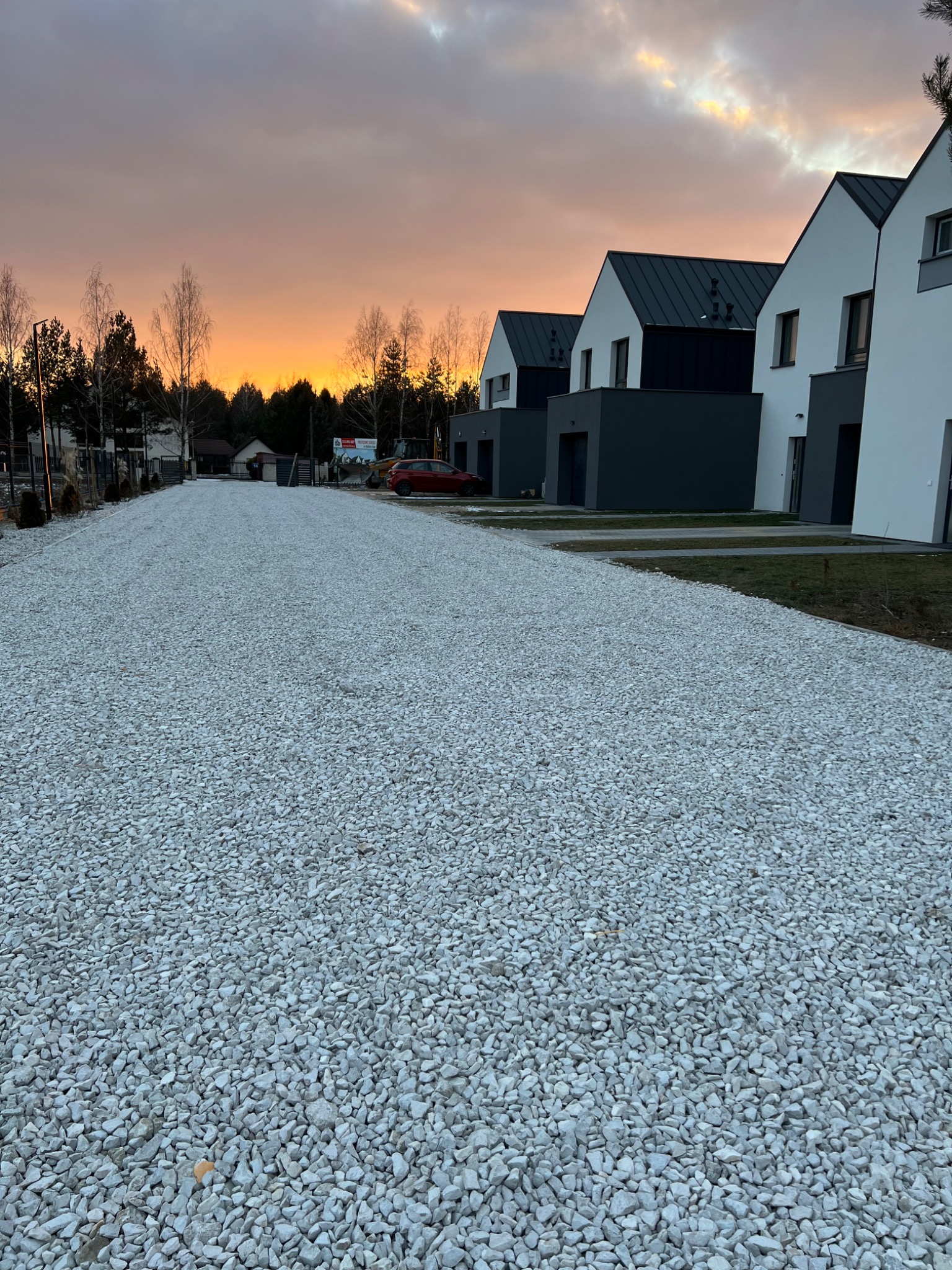 Utwardzona droga z białego kruszywa prowadząca do szeregu nowoczesnych domów w minimalistycznym stylu, tło stanowi zachodzące słońce i drzewa.