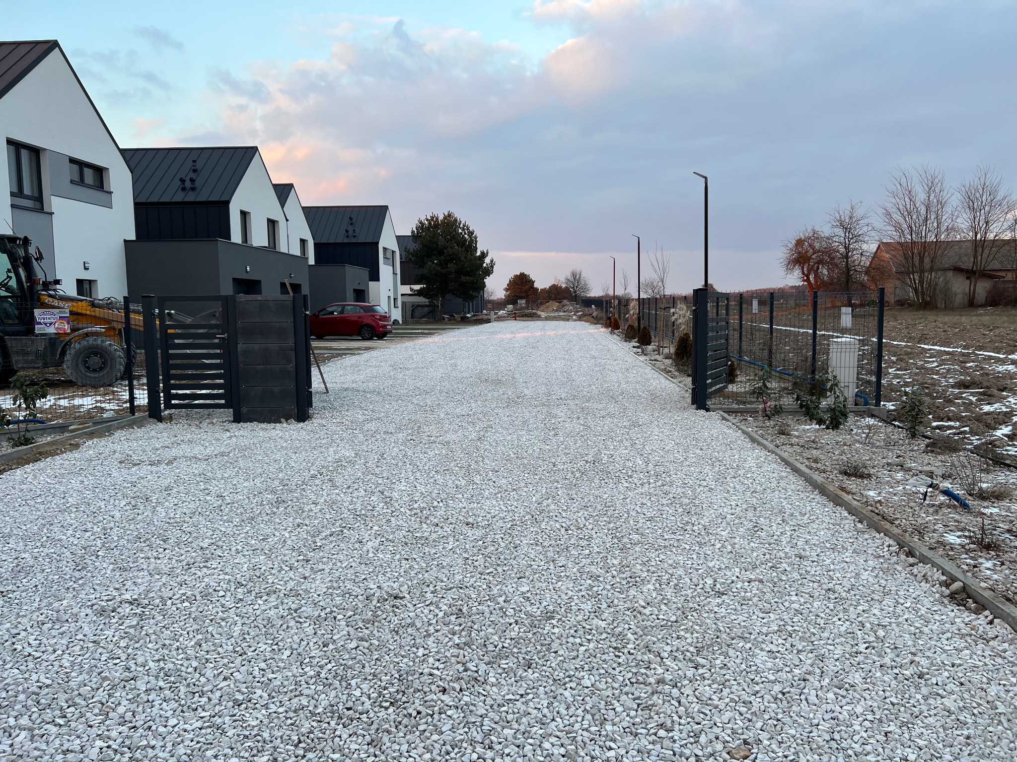 Biała, kamienista droga dojazdowa prowadząca do nowoczesnych domów jednorodzinnych, częściowo ogrodzona czarnym metalowym płotem, z widoczną koparką w tle.