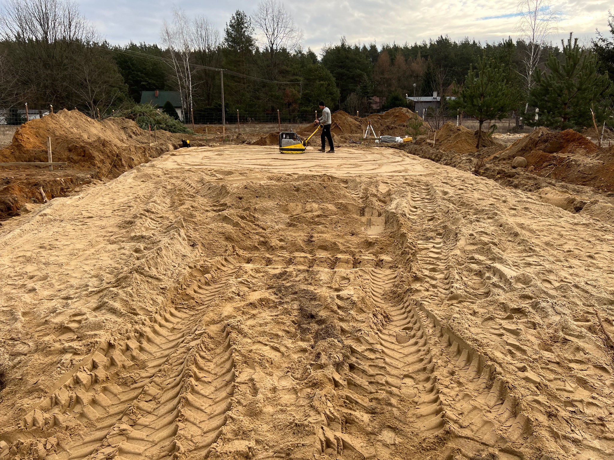 Praca zagęszczarką płytową na placu budowy, widoczne ślady opon na piasku, przygotowanie terenu pod budowę w otoczeniu drzew.