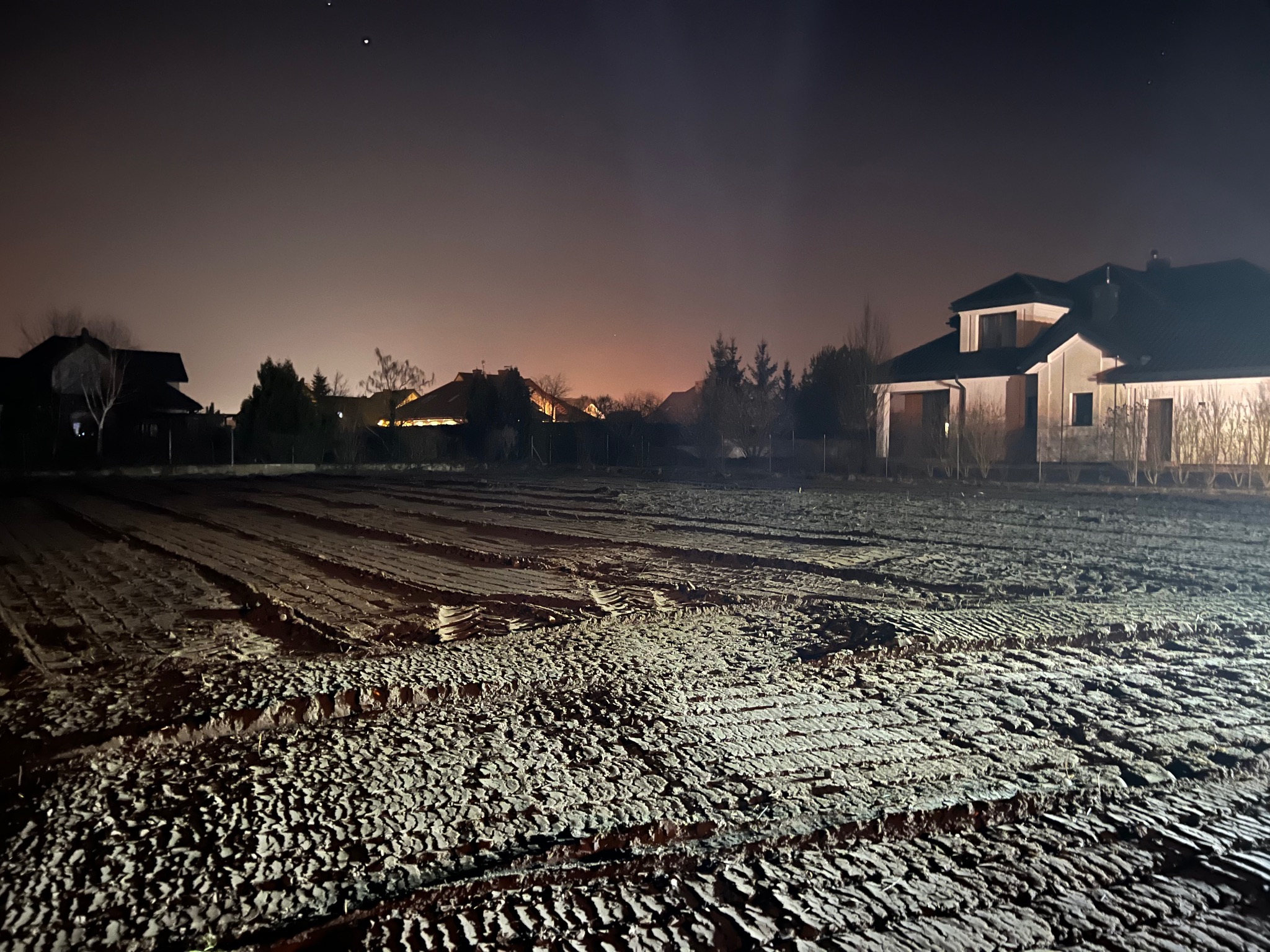 Przygotowany teren pod budowę w nocy, widoczne ślady po maszynach, w tle zabudowa jednorodzinna.