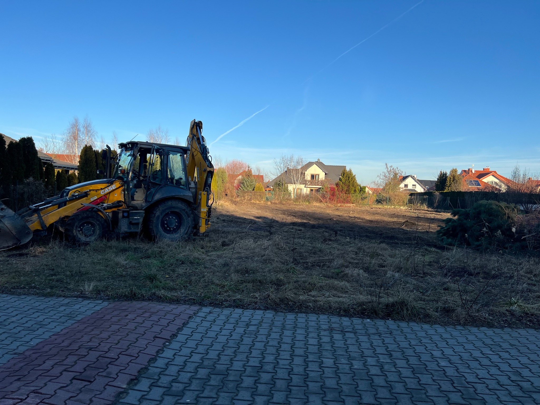Żółto-czarna koparko-ładowarka Case stoi na przygotowanym placu budowy z widokiem na domy jednorodzinne w tle, błękitne niebo.