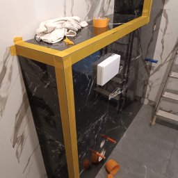 CEGIEŁKA Tomasz Goździela - Wykończona obudowa z czarnego marmuru w łazience, zabezpieczona żółtą taśmą malarską. Widoczne przyłącza wodne, biała zabudowa i drabina. Glazura podłogowa w odcieniach szarości.