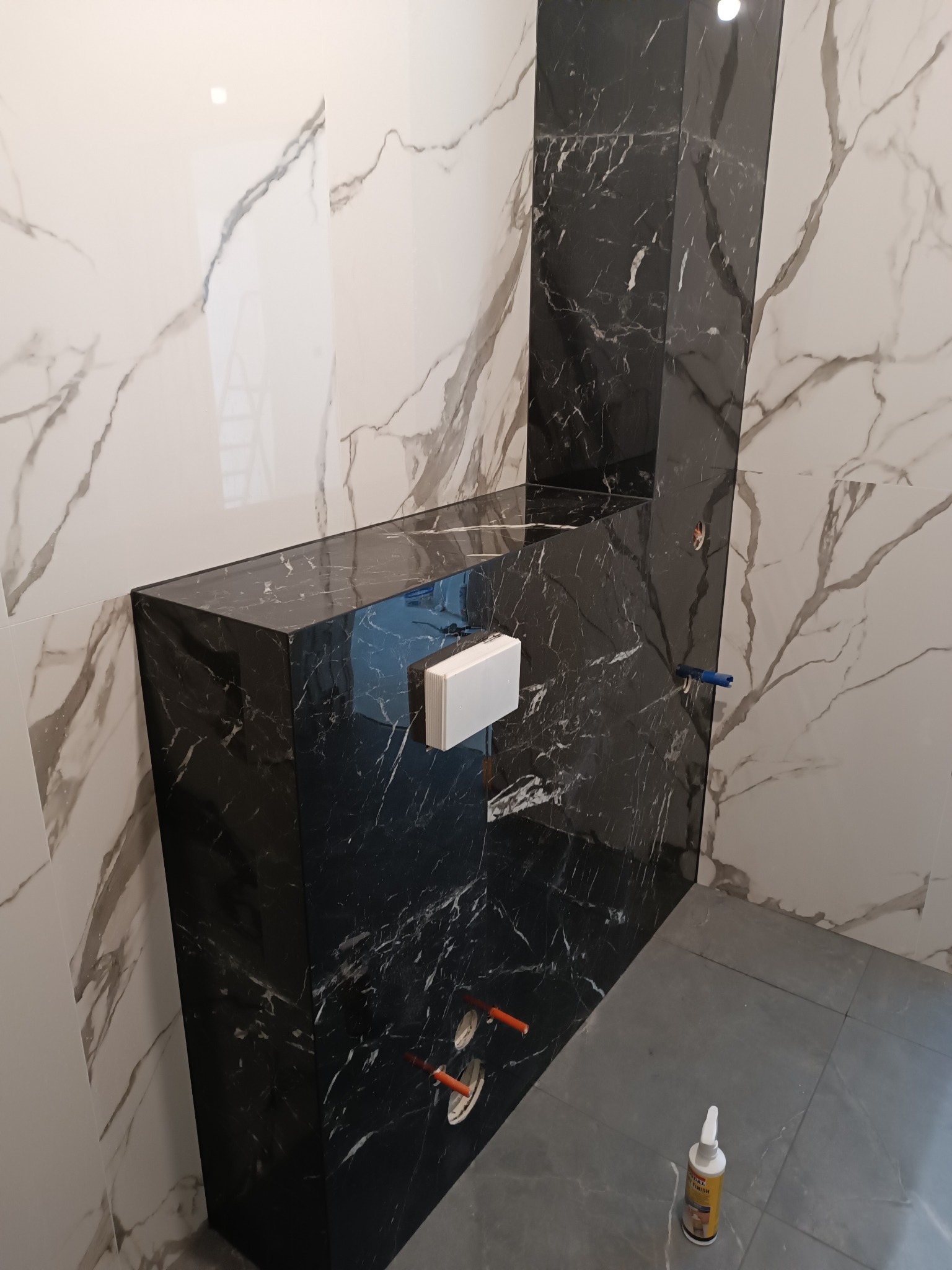 Wykończona łazienka z czarno-białymi płytkami imitującymi marmur. Widoczna zabudowa stelaża WC z czarnych płyt. Na podłodze szare płytki. Użyto środka do czyszczenia.