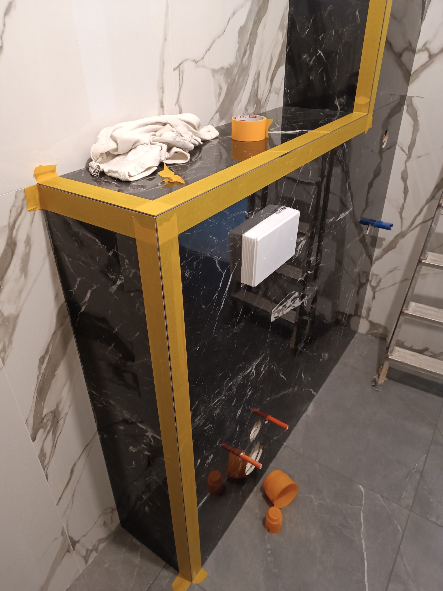 Wykończona obudowa z czarnego marmuru w łazience, zabezpieczona żółtą taśmą malarską. Widoczne przyłącza wodne, biała zabudowa i drabina. Glazura podłogowa w odcieniach szarości.