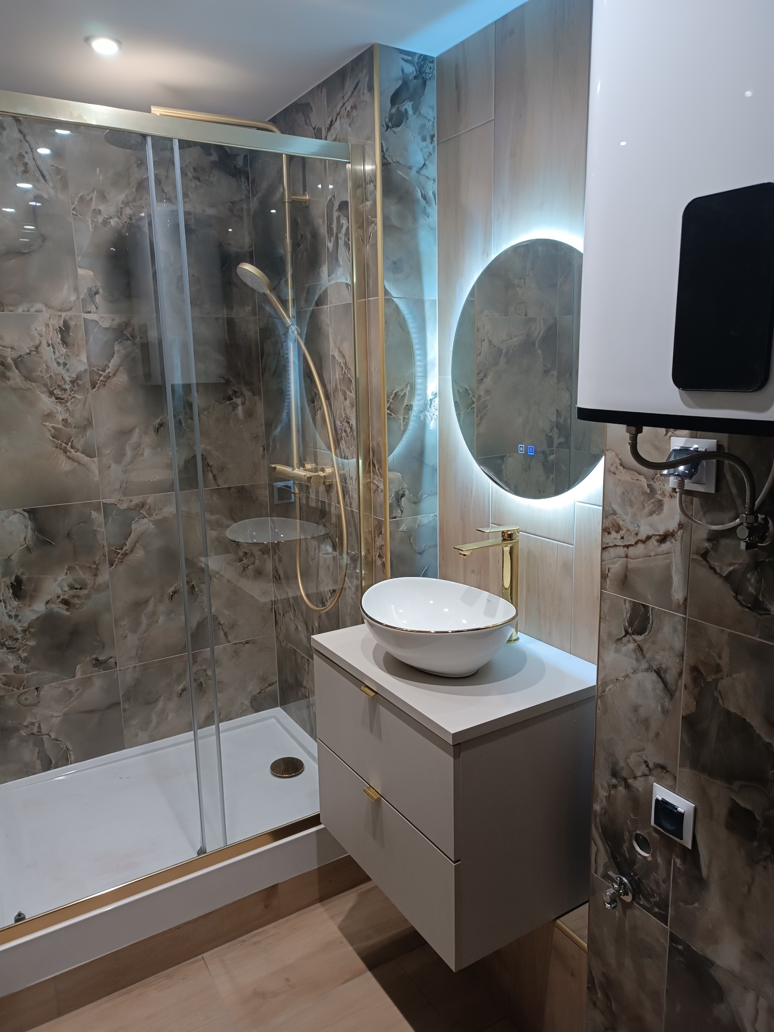 Nowoczesna łazienka z kabiną prysznicową typu walk-in, umywalką nablatową na szafce i okrągłym lustrem z podświetleniem LED. Płytki imitujące marmur.