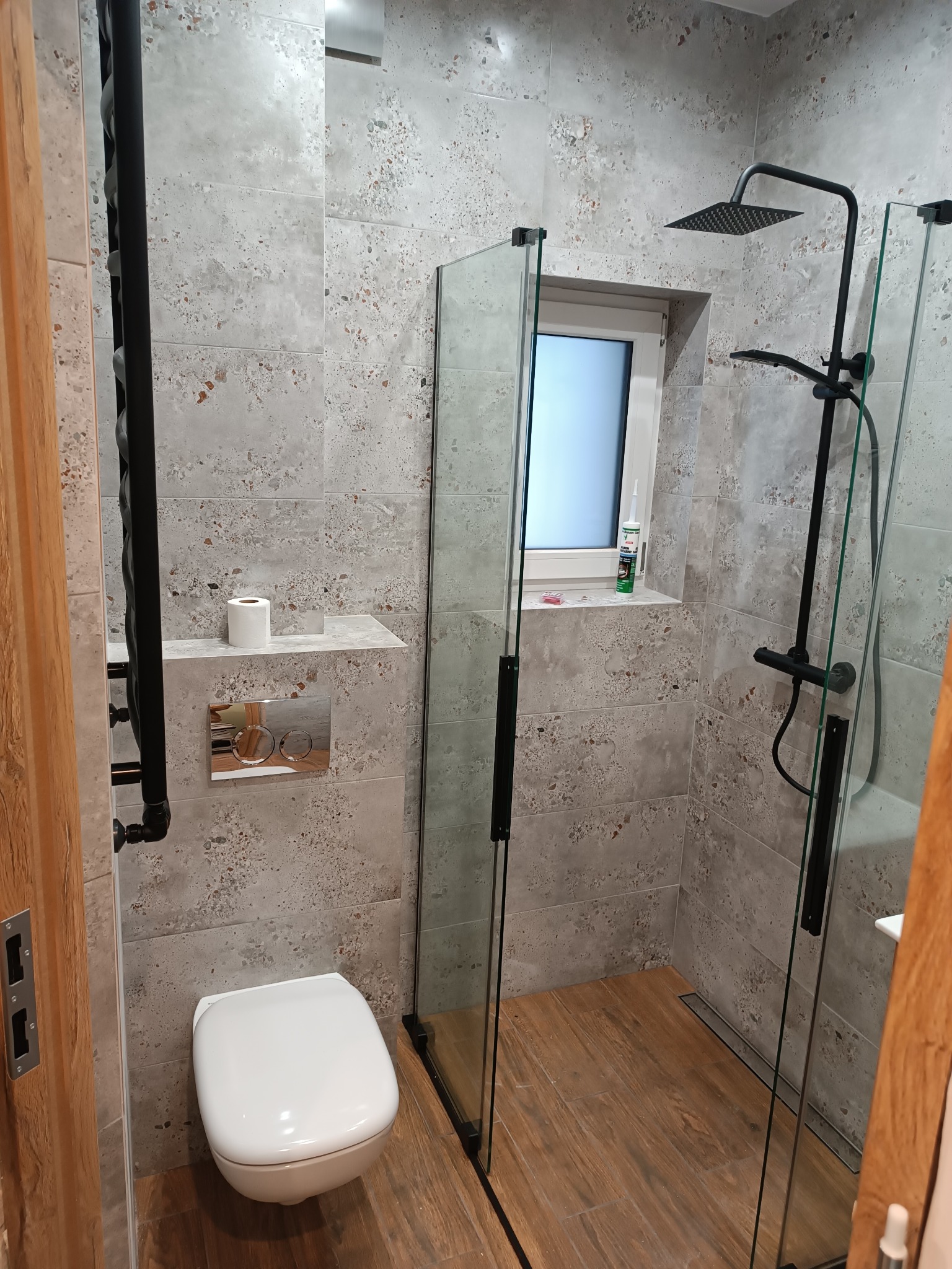 Nowoczesna łazienka z kabiną typu walk-in, szare płytki imitujące kamień, drewnopodobna podłoga, czarna armatura i grzejnik drabinkowy. Minimalistyczny design.