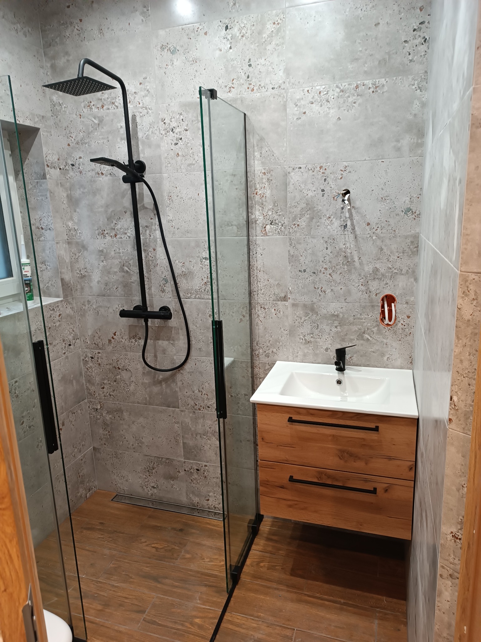 Nowoczesna łazienka z kabiną typu walk-in, czarną armaturą i drewnianą szafką pod umywalką. Płytki imitujące kamień na ścianach i drewnopodobne na podłodze.