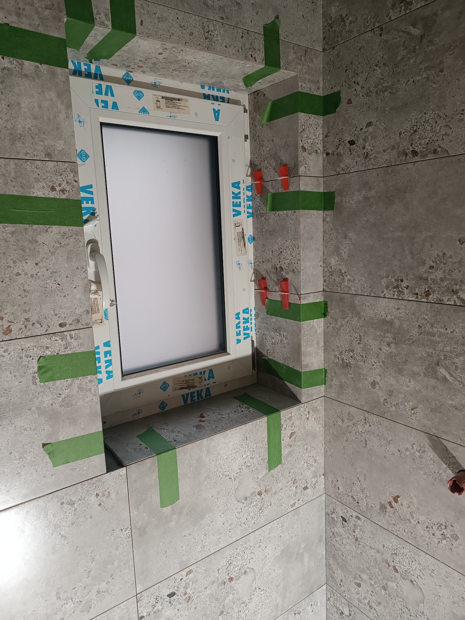 Okładzina z płytek imitujących lastryko wokół okna, zabezpieczona taśmą malarską i klinami poziomującymi w Paczkowie. Widoczne detale wykończenia.