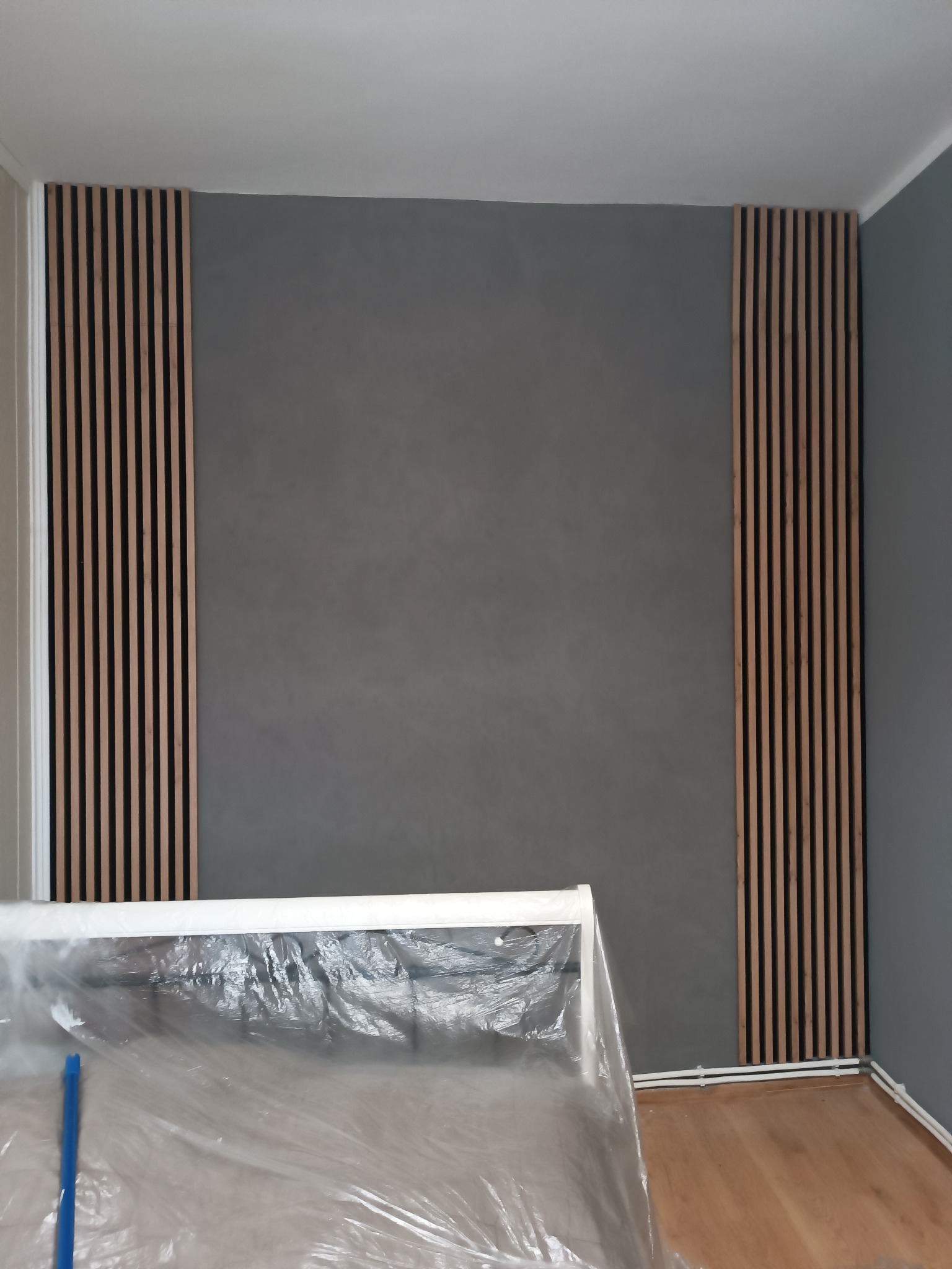 Szara ściana z dekoracyjnymi, drewnianymi lamelami po bokach. Podłoga zabezpieczona folią malarską. Minimalistyczny design wnętrza po remoncie.