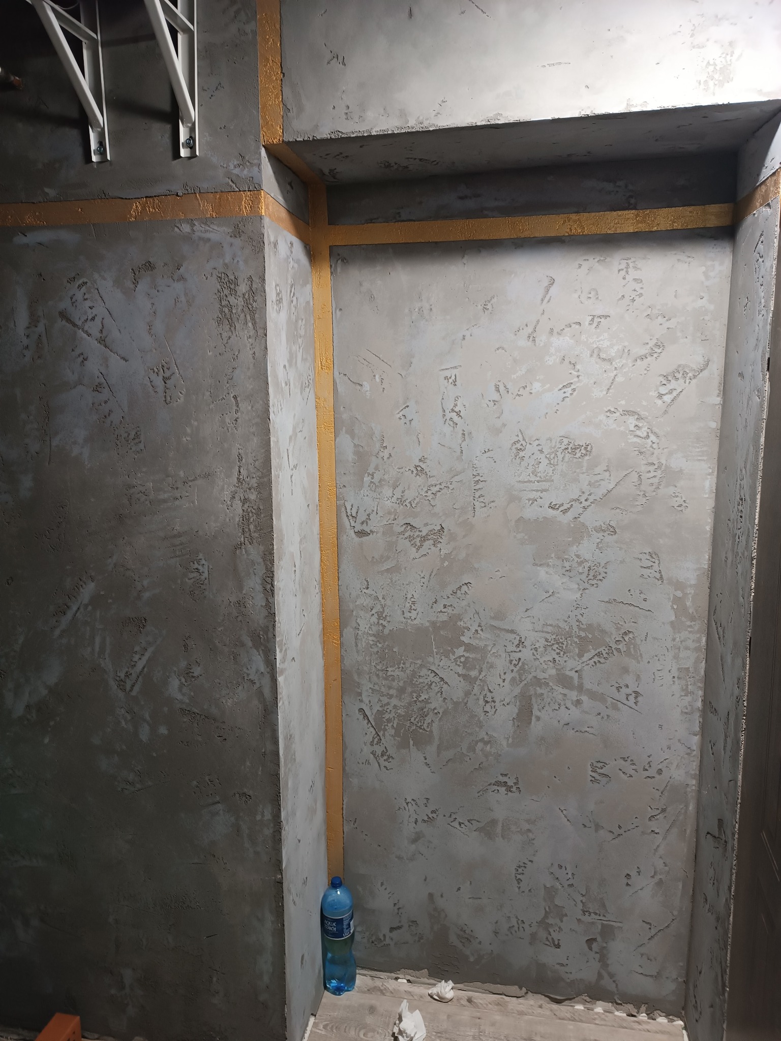 Ściana z szarą strukturą, oddzielona złotą taśmą malarską. Widoczna butelka wody i resztki materiałów budowlanych. Etap prac wykończeniowych w pomieszczeniu.